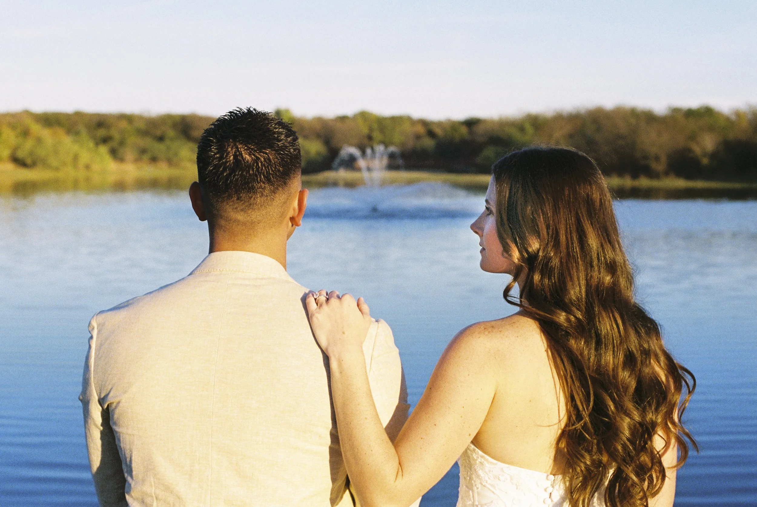 A couple by a lake, the woman with long wavy brown hair and a white dress, the man with short dark hair and a light-colored suit, standing close together with the woman resting her hand on the man's shoulder, surrounded by a calm body of water and a 