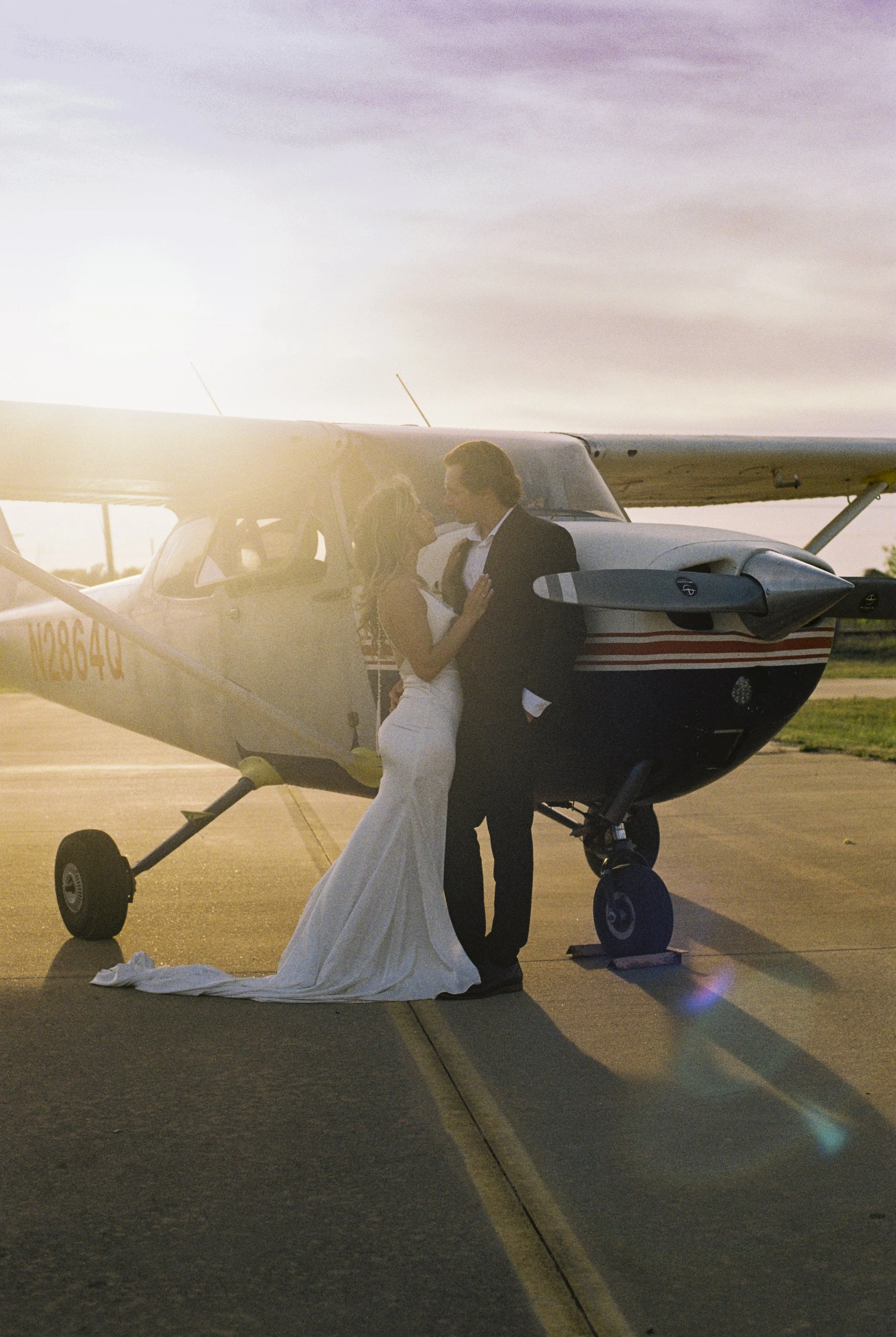 A couple in wedding attire stands next to a small airplane, holding hands and looking into each other's eyes during sunset on an airstrip.