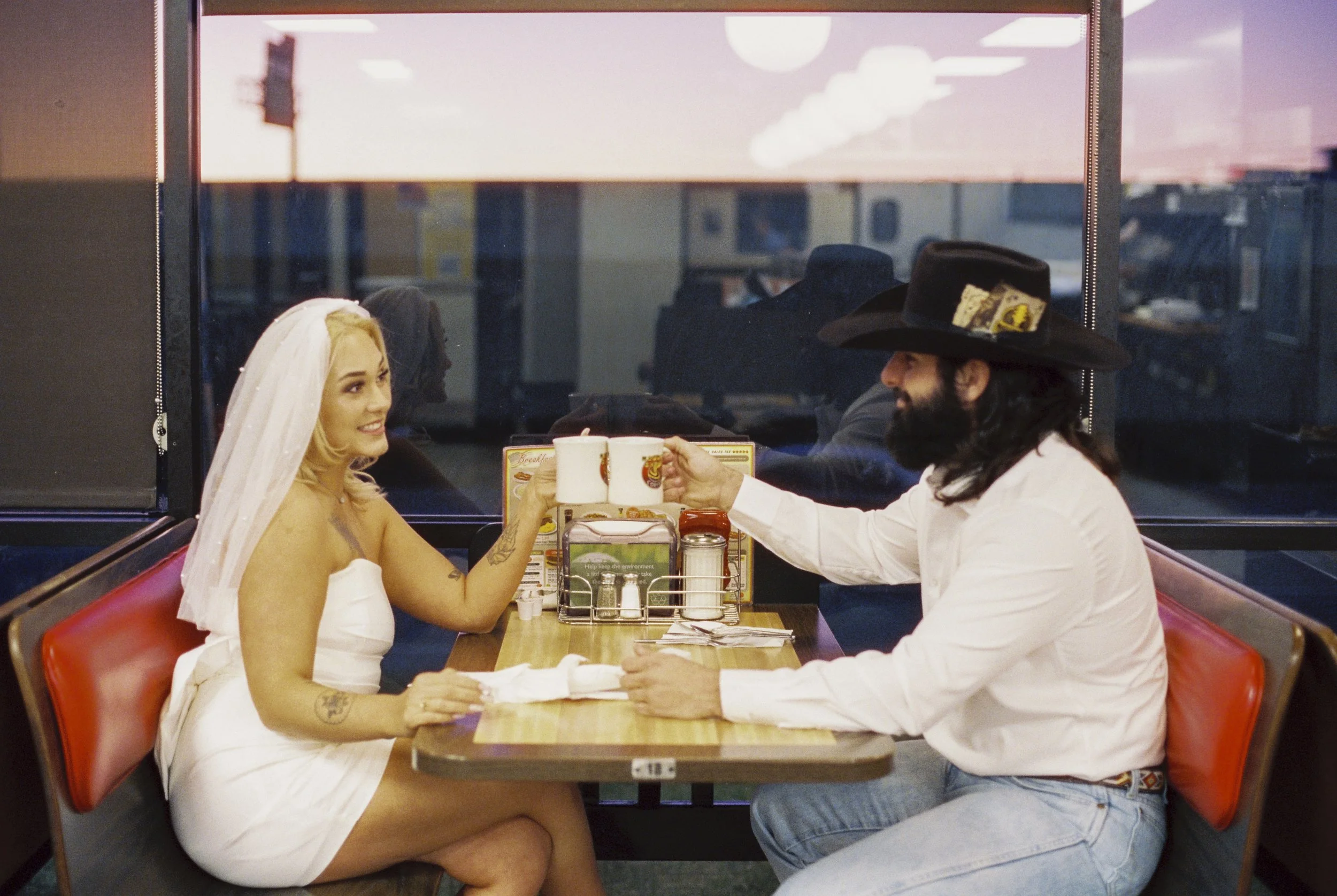 A man and woman sharing drinks at a diner. The woman is wearing a white dress and veil, and the man is wearing a white shirt, jeans, and a wide-brimmed black hat. They are smiling and clinking cups together in a friendly toast.
