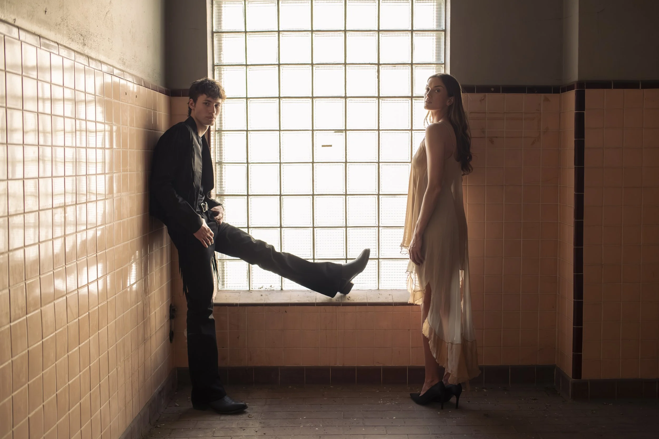 A young man and woman standing apart in a room with large glass block windows. The man, dressed in black with one leg resting on a ledge, looks toward the camera. The woman, wearing a light-colored dress, faces sideways and looks into the distance. T