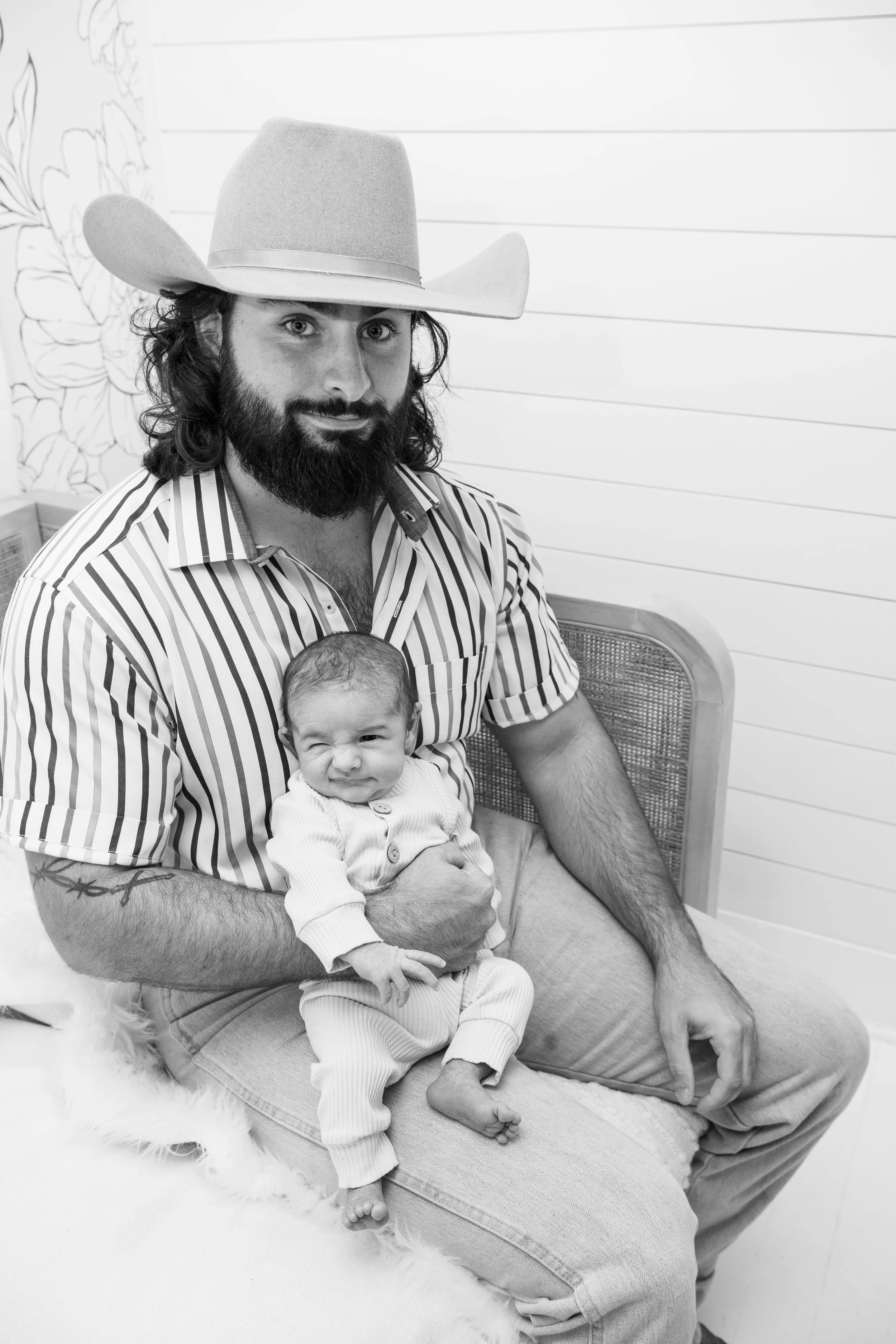 A man with a beard, long hair, striped shirt, and cowboy hat holds a wrinkling baby dressed in a light-colored outfit, sitting on his lap; they are in a cozy room with a wooden wall and floral wallpaper background.