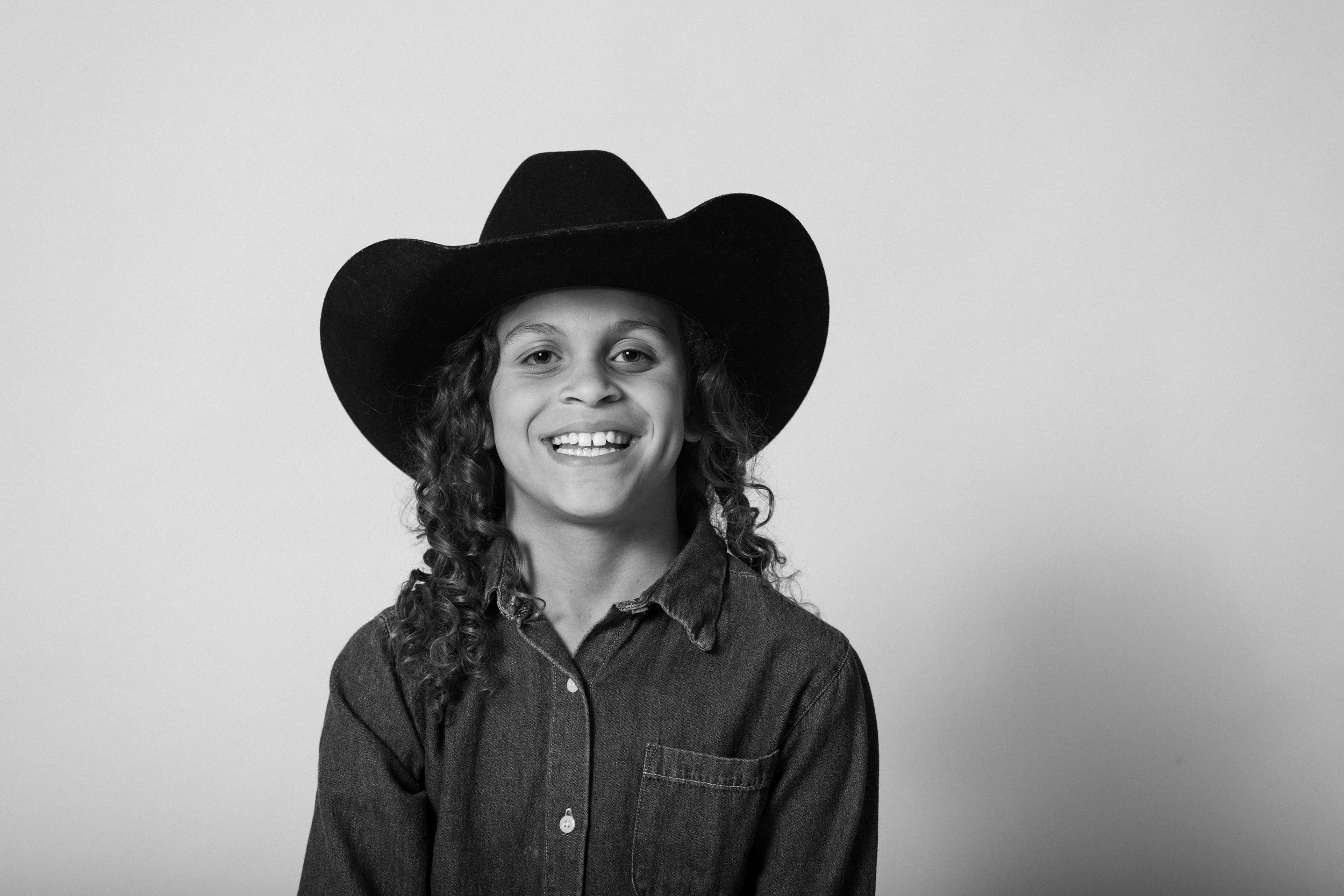 A boy with curly hair wearing a cowboy hat and denim shirt, smiling at the camera.