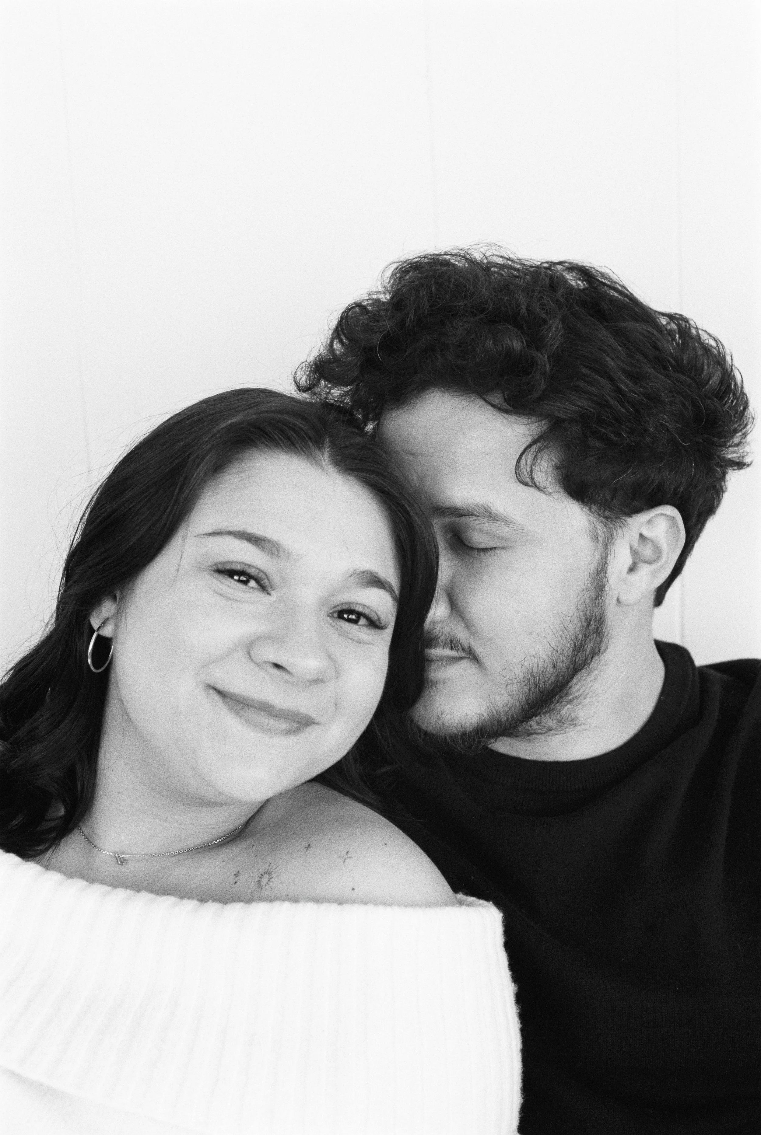 A black and white photo of a smiling woman with dark hair and hoop earrings, and a man with curly hair and a beard, with his eyes closed, leaning in close to her.