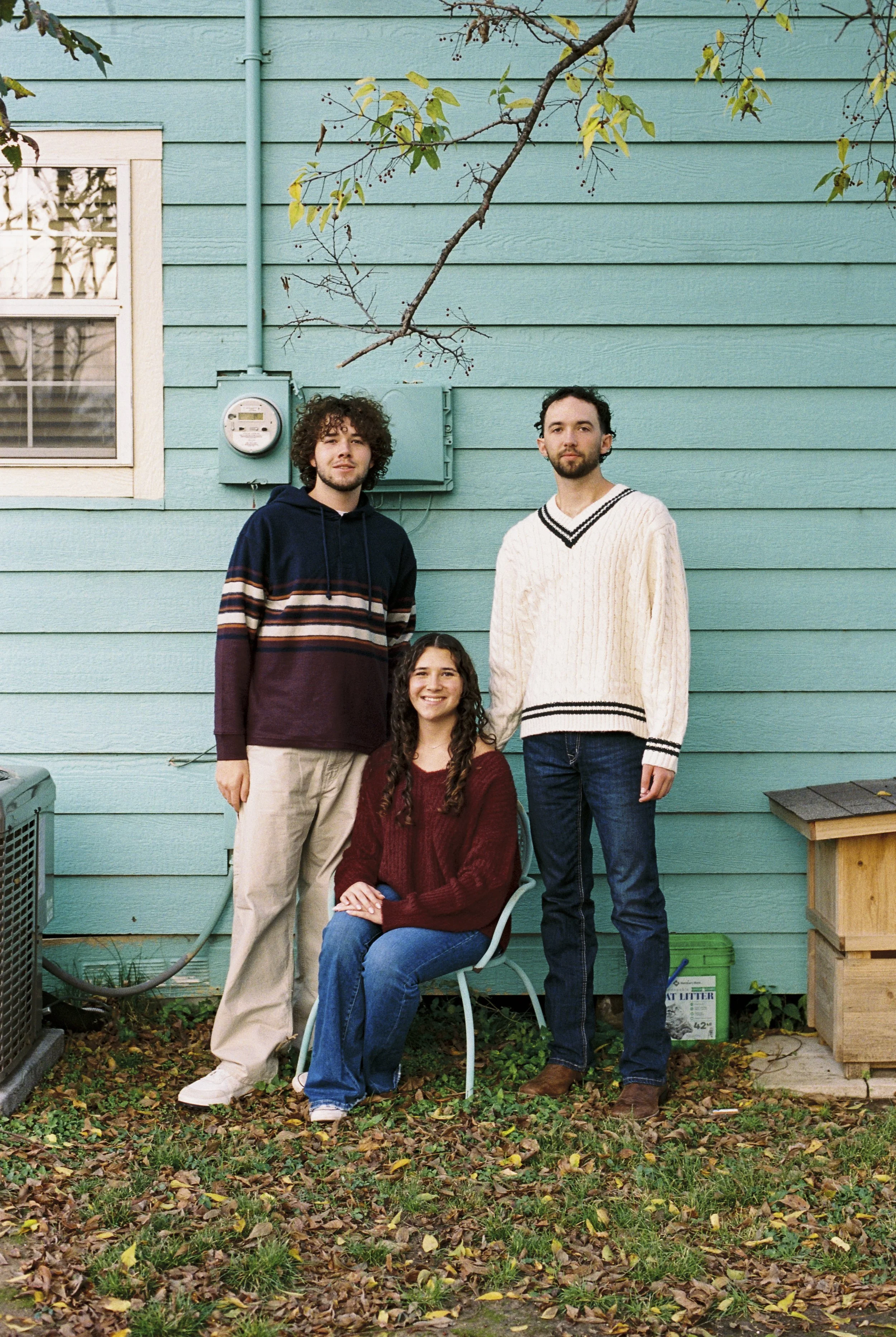Three people outdoors in front of a turquoise house wall, standing or sitting on a chair. One woman is sitting in the center with long curly hair, wearing a maroon sweater and blue jeans. Two men are standing on either side; one with curly hair in a 