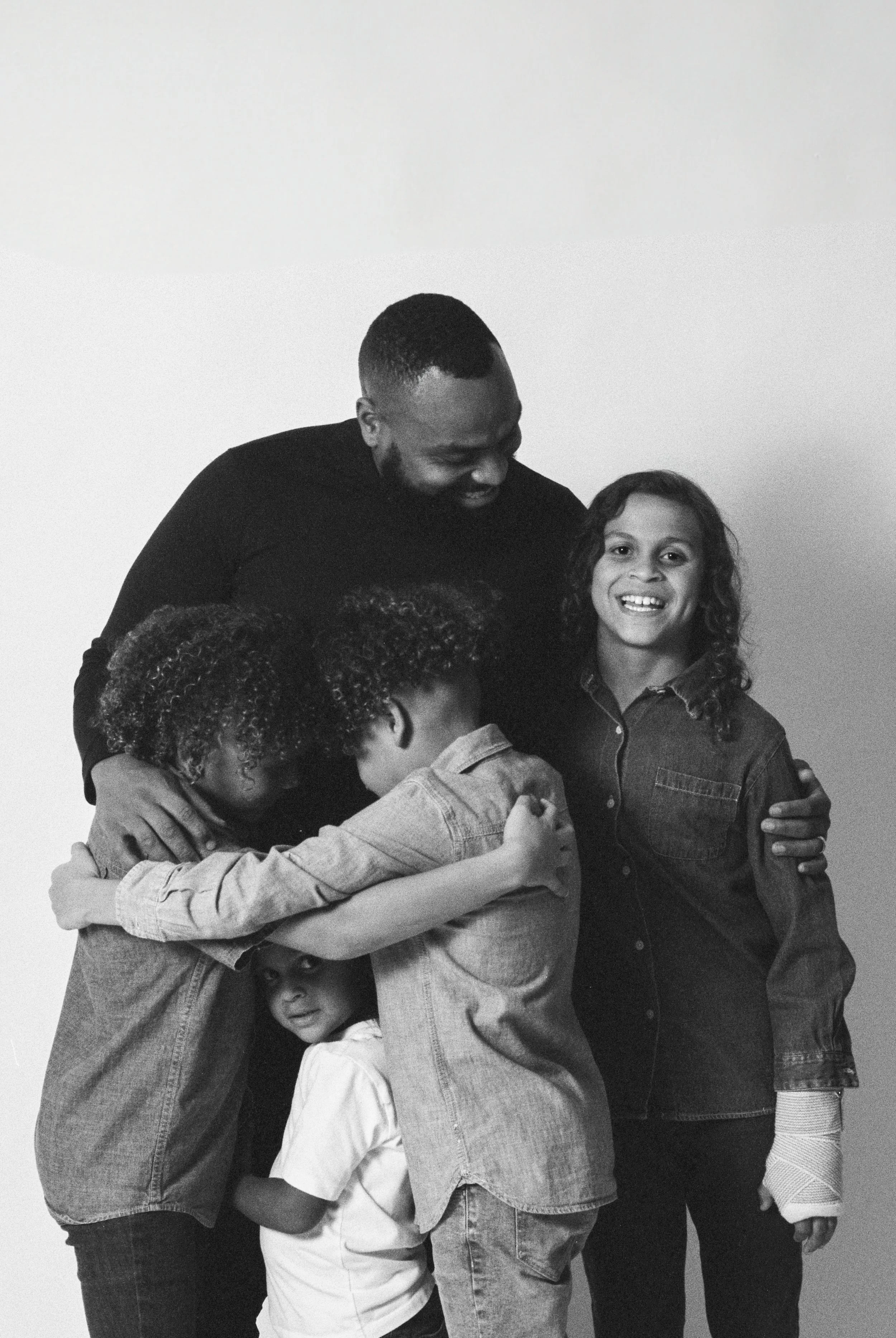 A black-and-white photo of a family of five happily hugging against a plain white background. The family consists of a man and four children, all smiling and embracing each other.