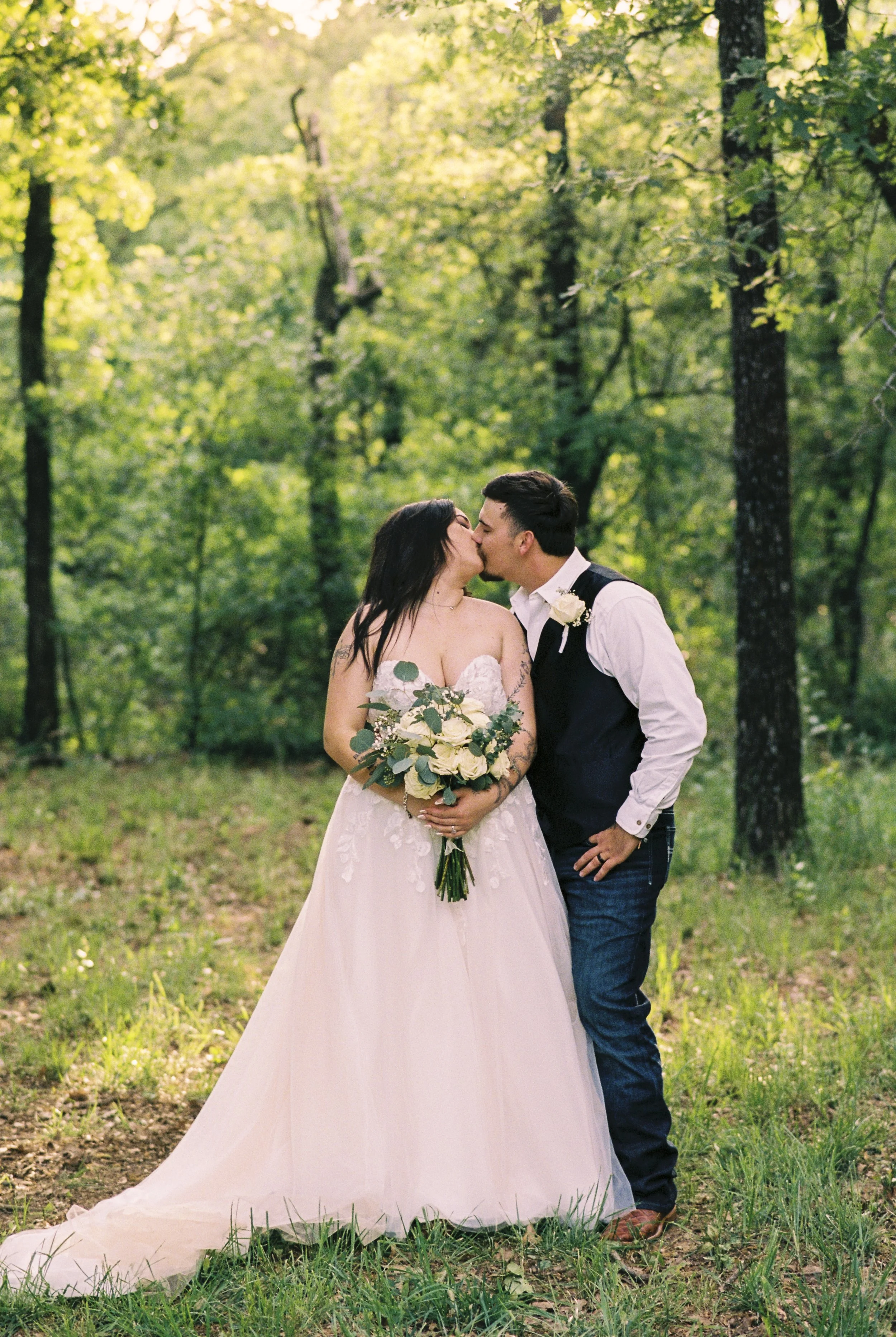 A wedding couple is kissing in a lush green forest, with the bride holding a bouquet of white roses and greenery, dressed in a white wedding gown and the groom in a white shirt, black vest, and jeans.