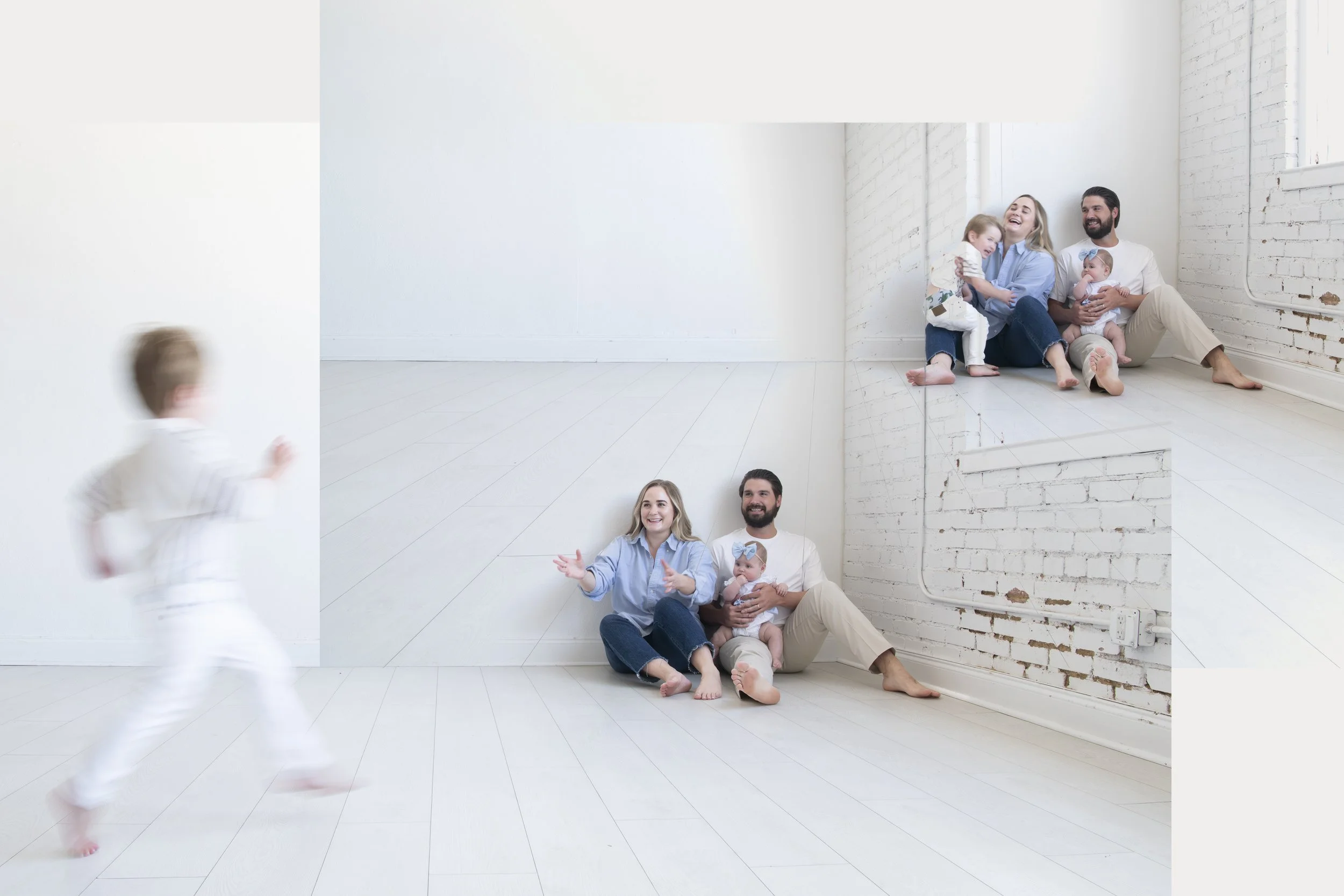 A family of four sitting on the floor in front of a mirror, with a young child running past. The family is smiling and enjoying a playful moment at home with a white brick wall and large windows in the background.