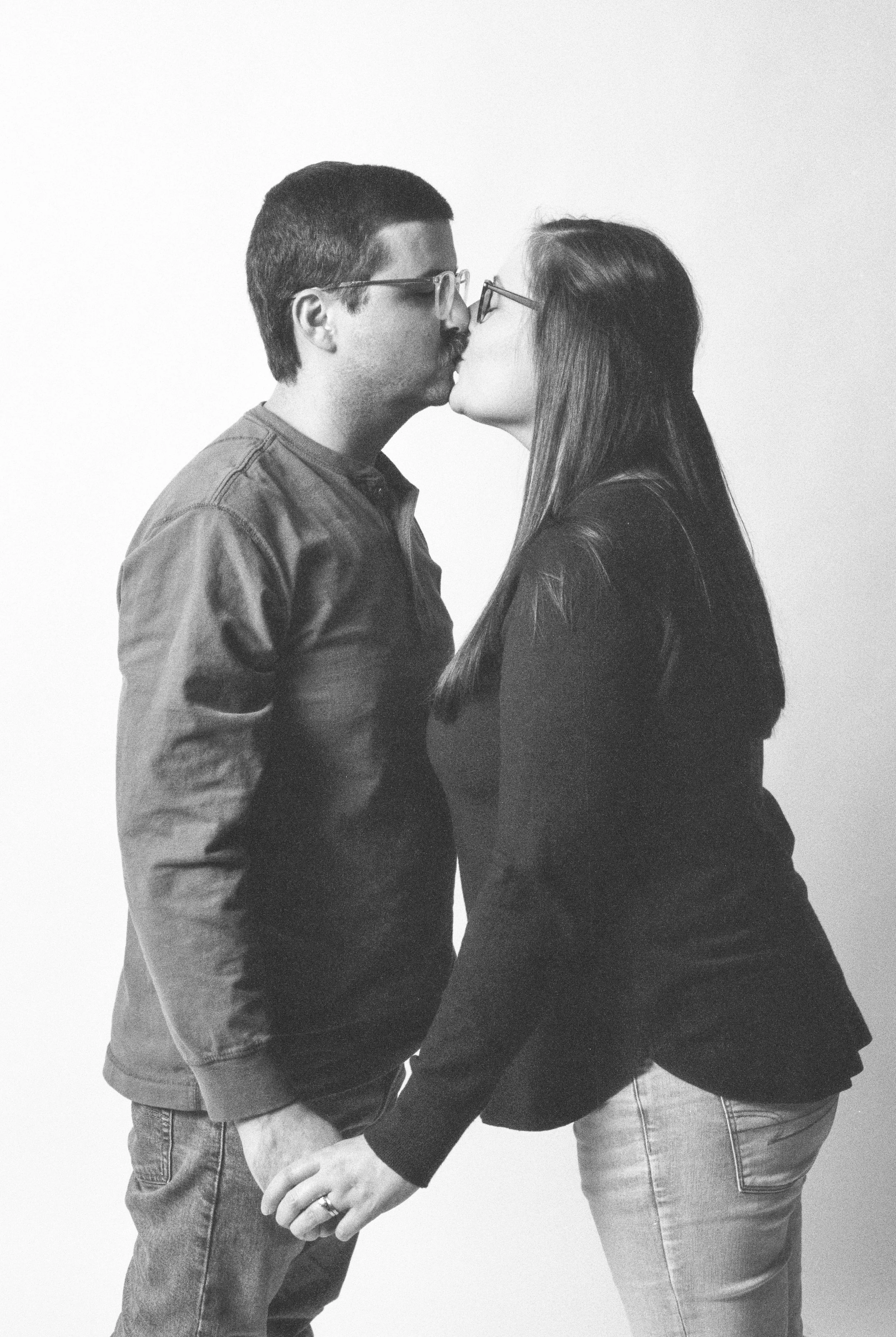 A black-and-white photo of a man and woman kissing, holding hands, with glasses, standing against a plain wall.