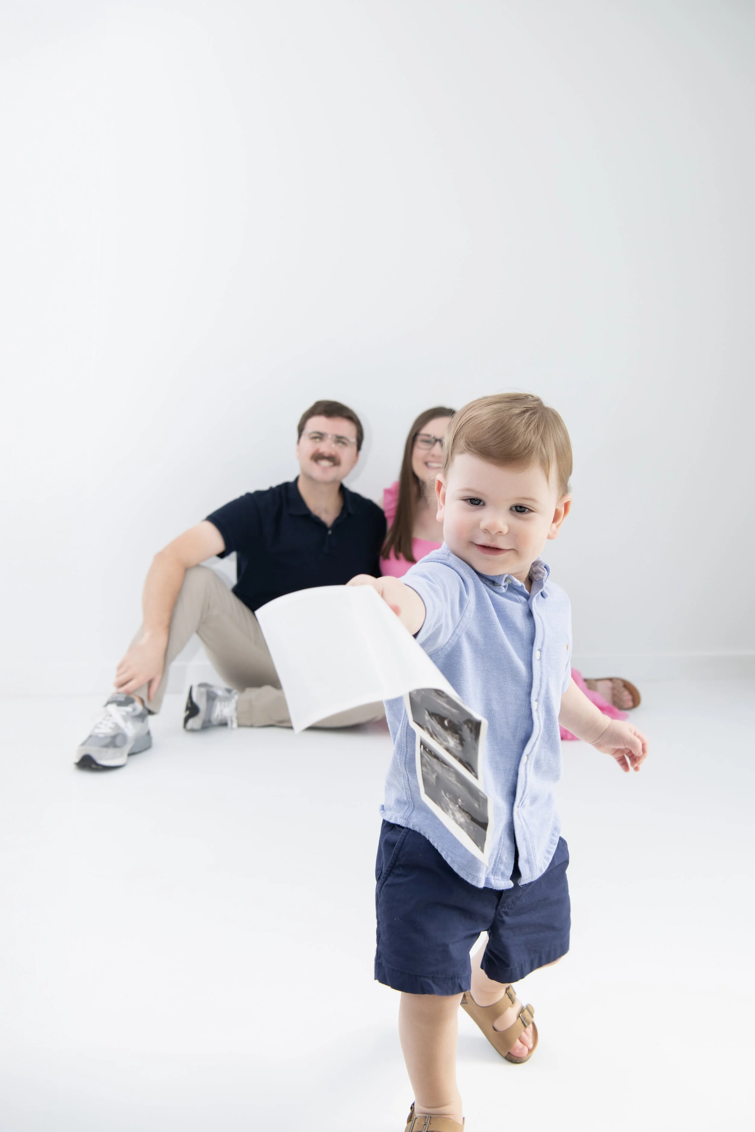 Young boy in blue shirt and shorts holding ultrasound pictures, with a smiling couple sitting on the floor behind him.