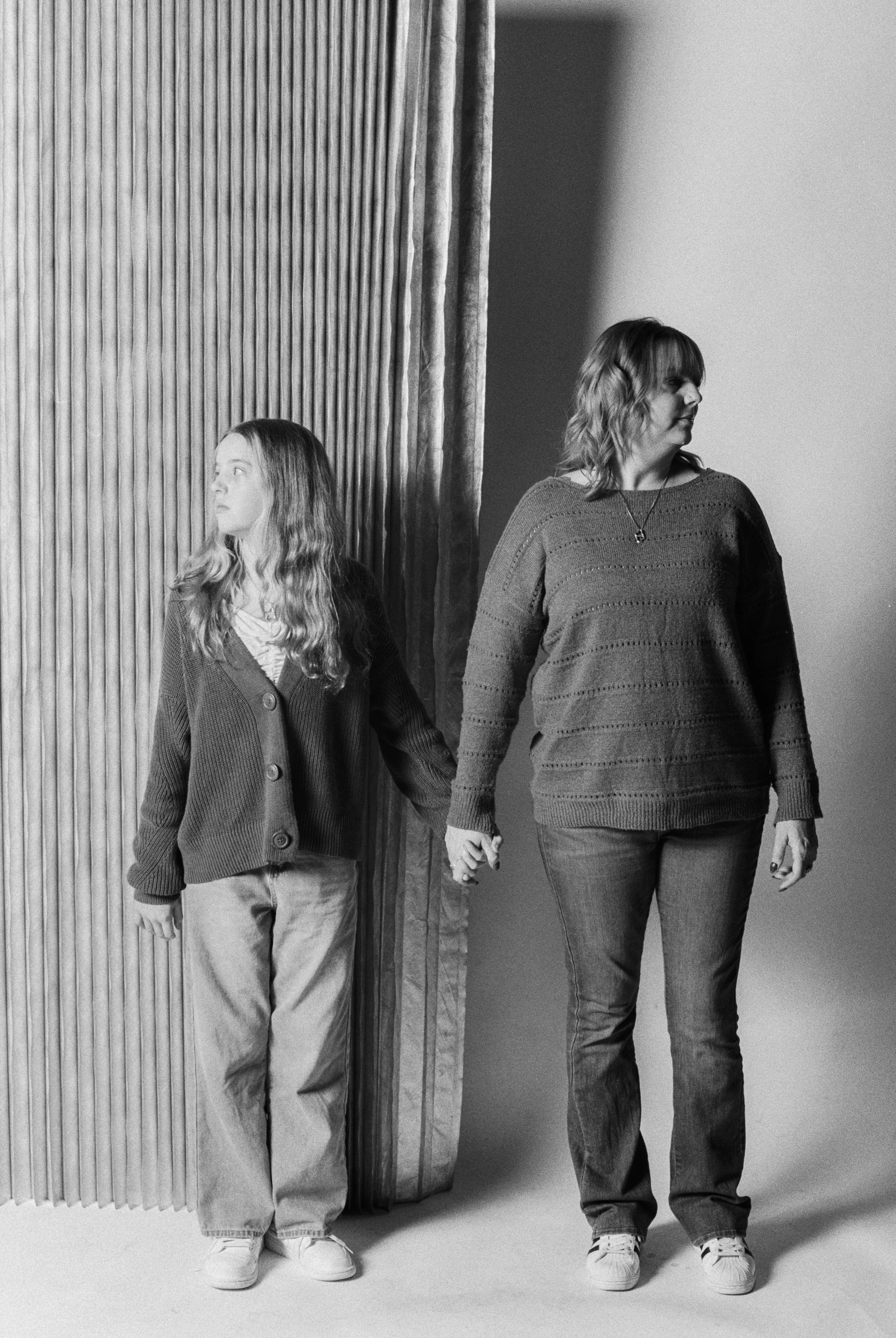 A woman and a girl holding hands, looking away in a black and white photo, standing in front of a textured curtain.