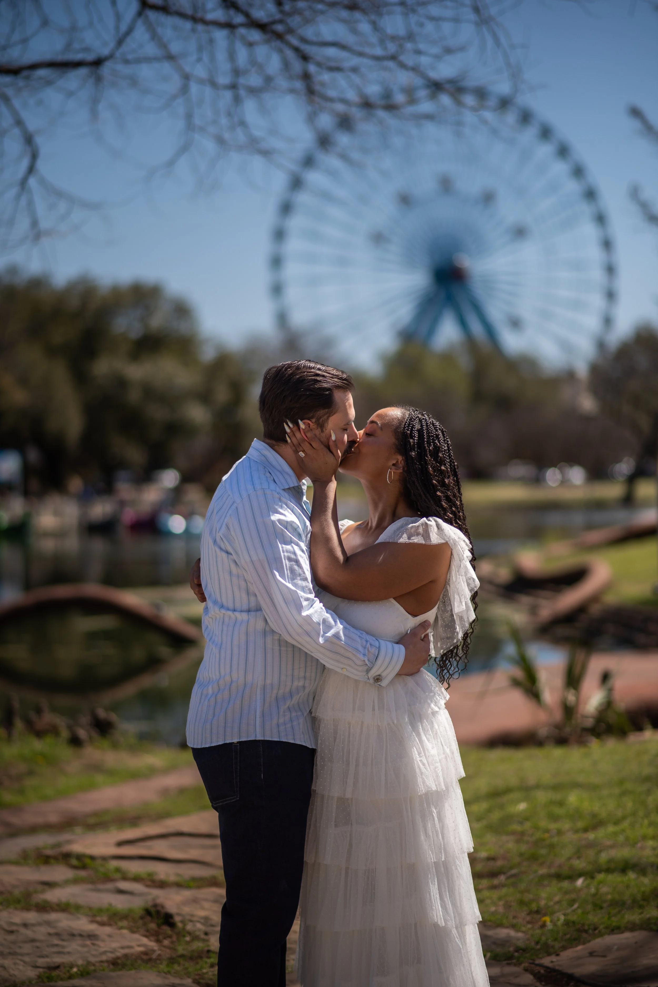 A couple sharing a kiss outdoors near a pond with a Ferris wheel in the background on a sunny day.