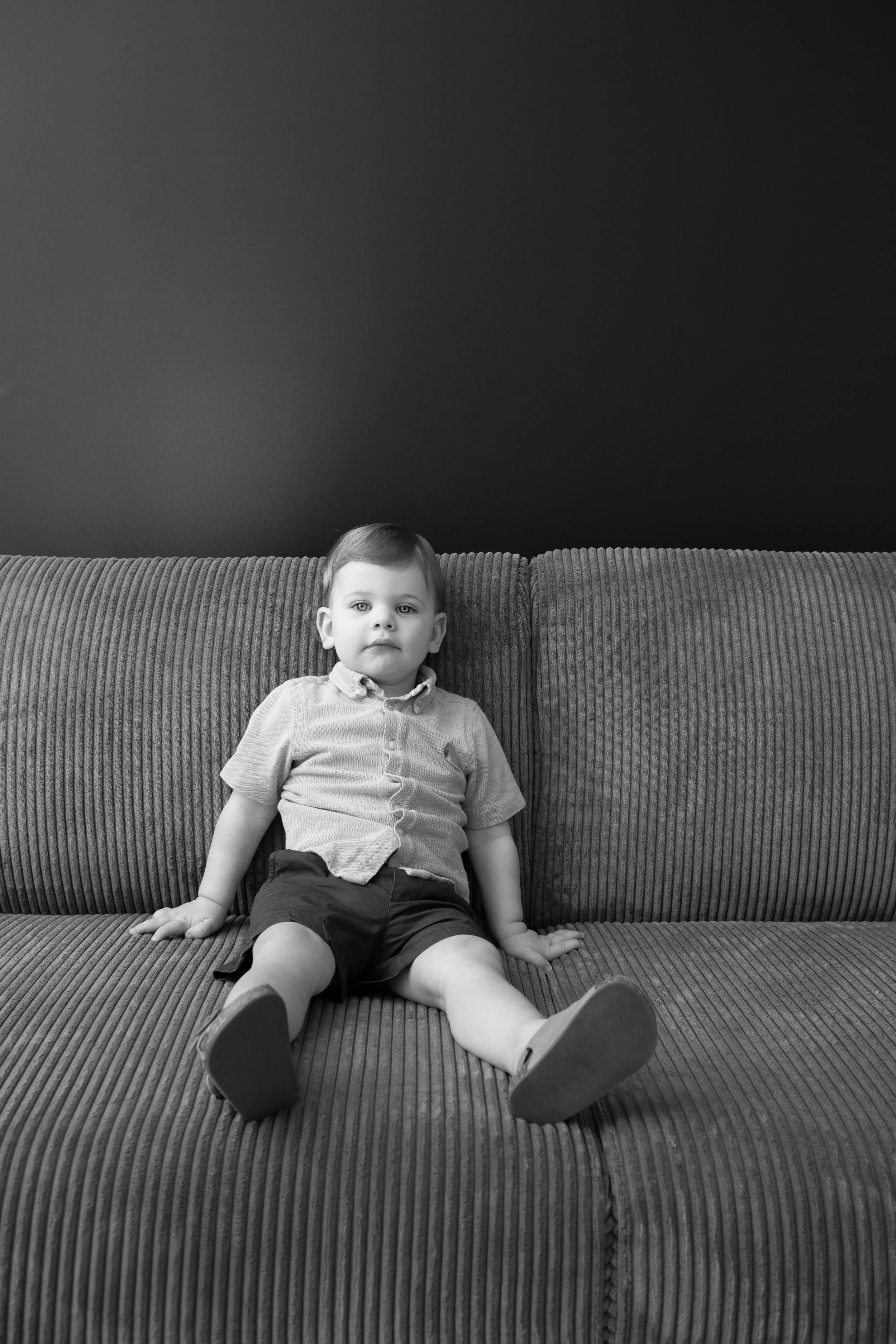 A young boy sitting on a ribbed fabric sofa, looking at the camera, against a plain dark wall.