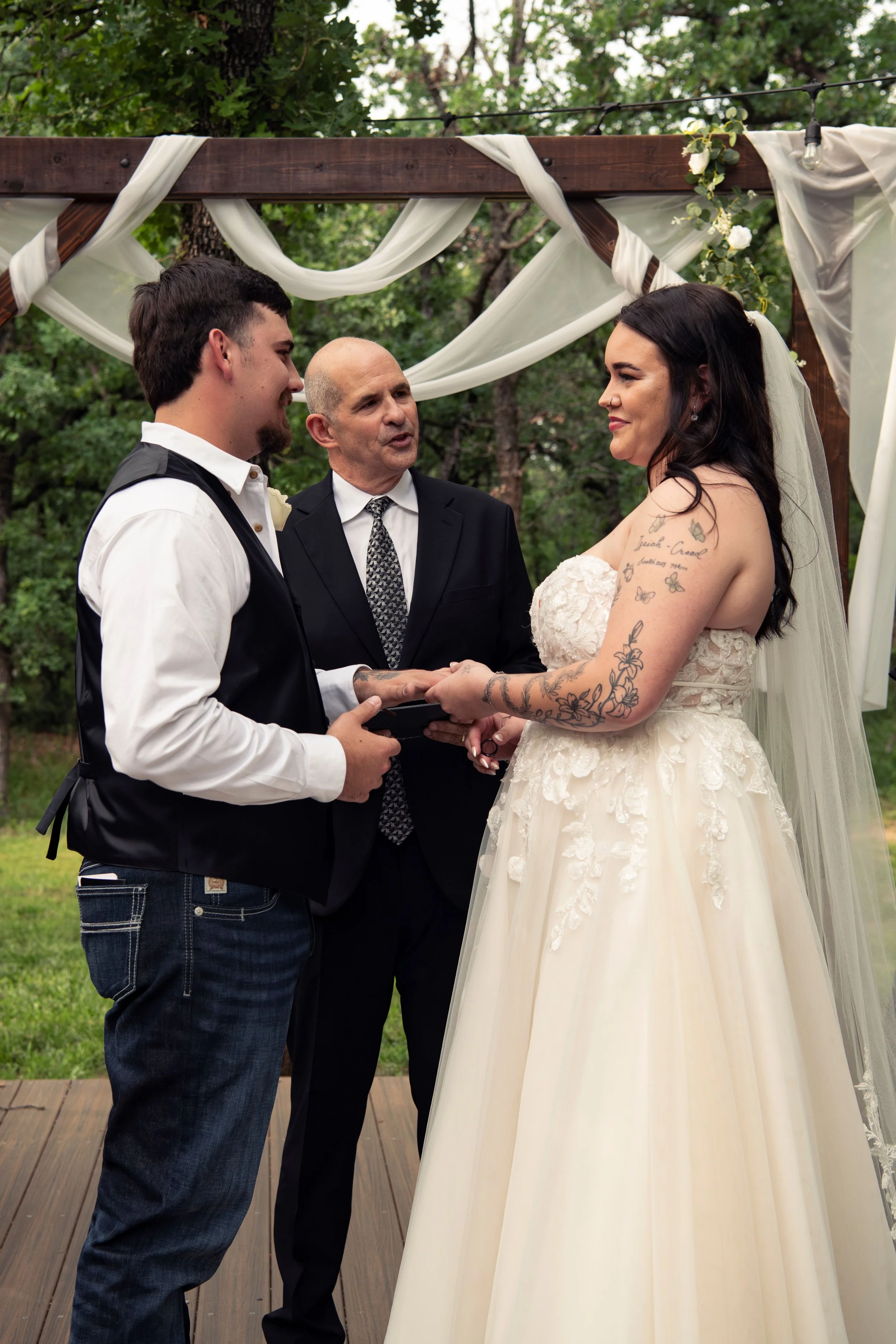 A wedding ceremony with a bride and groom holding hands, facing an officiant under a decorated wooden arch outdoors surrounded by trees.