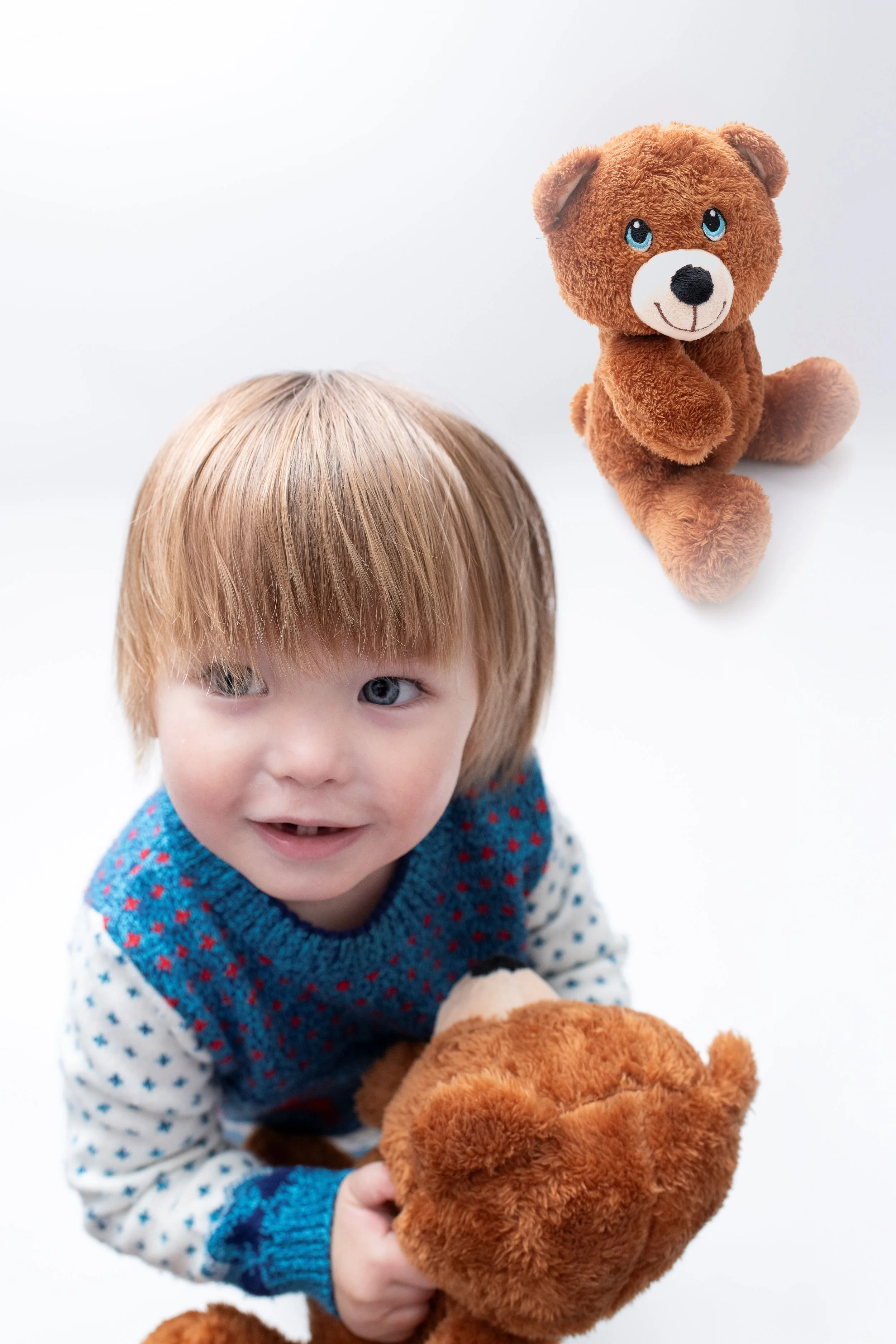 A young child with blond hair and blue eyes holding a brown teddy bear, with another teddy bear on the white background.