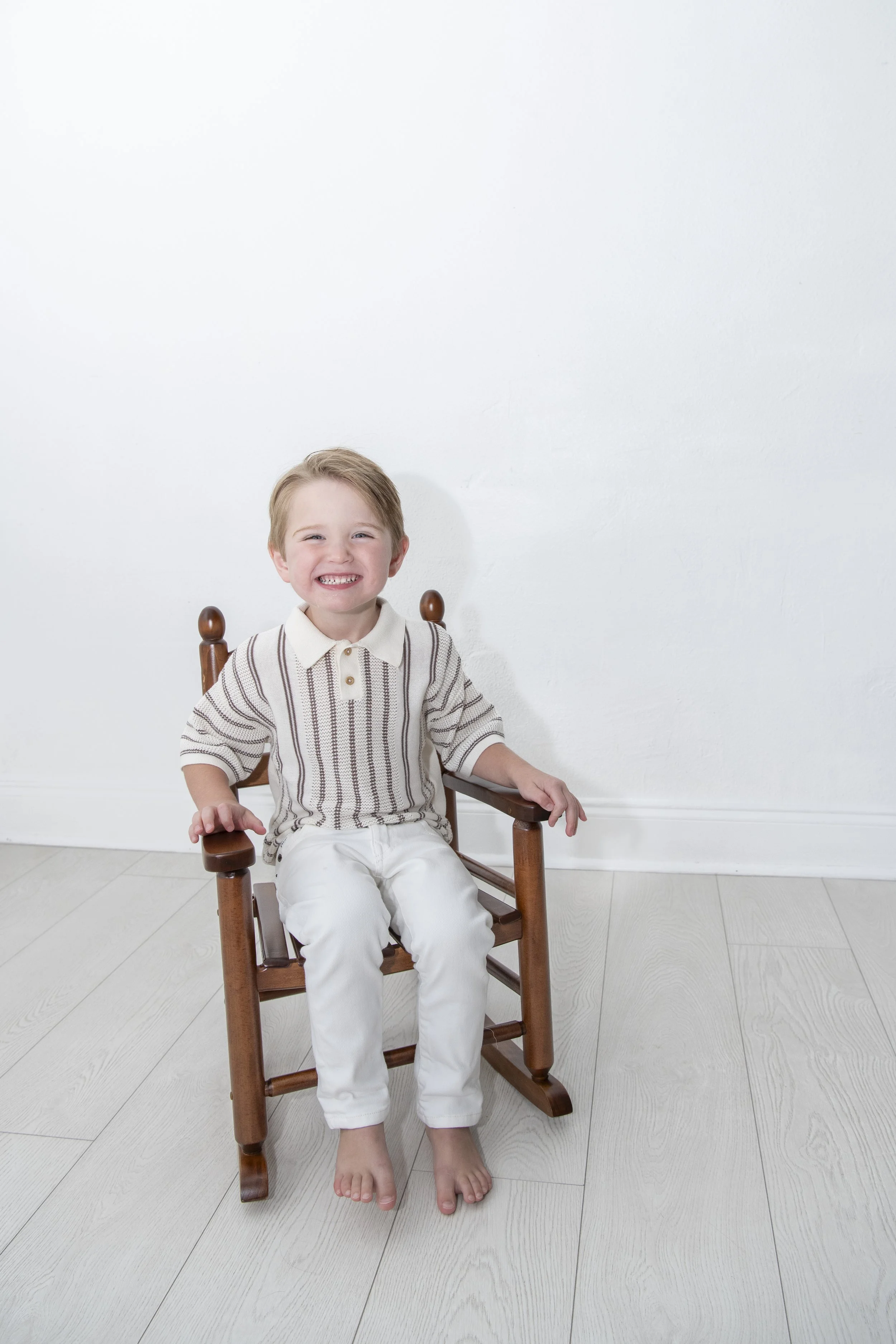 A young boy with light hair and a big smile sitting in a wooden chair against a plain white wall, wearing a striped shirt and white pants.