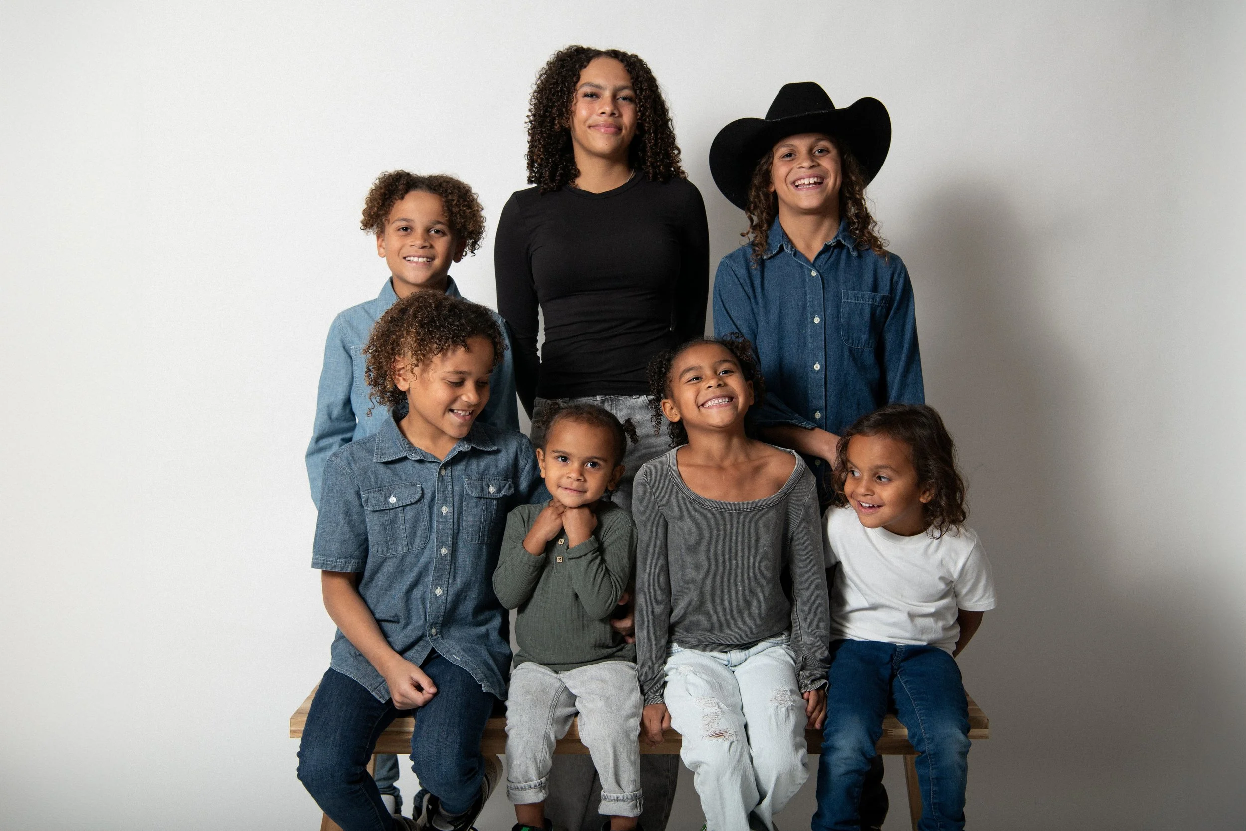 A group of seven diverse children posing together against a plain background, smiling and wearing casual clothing, with some children sitting and others standing.