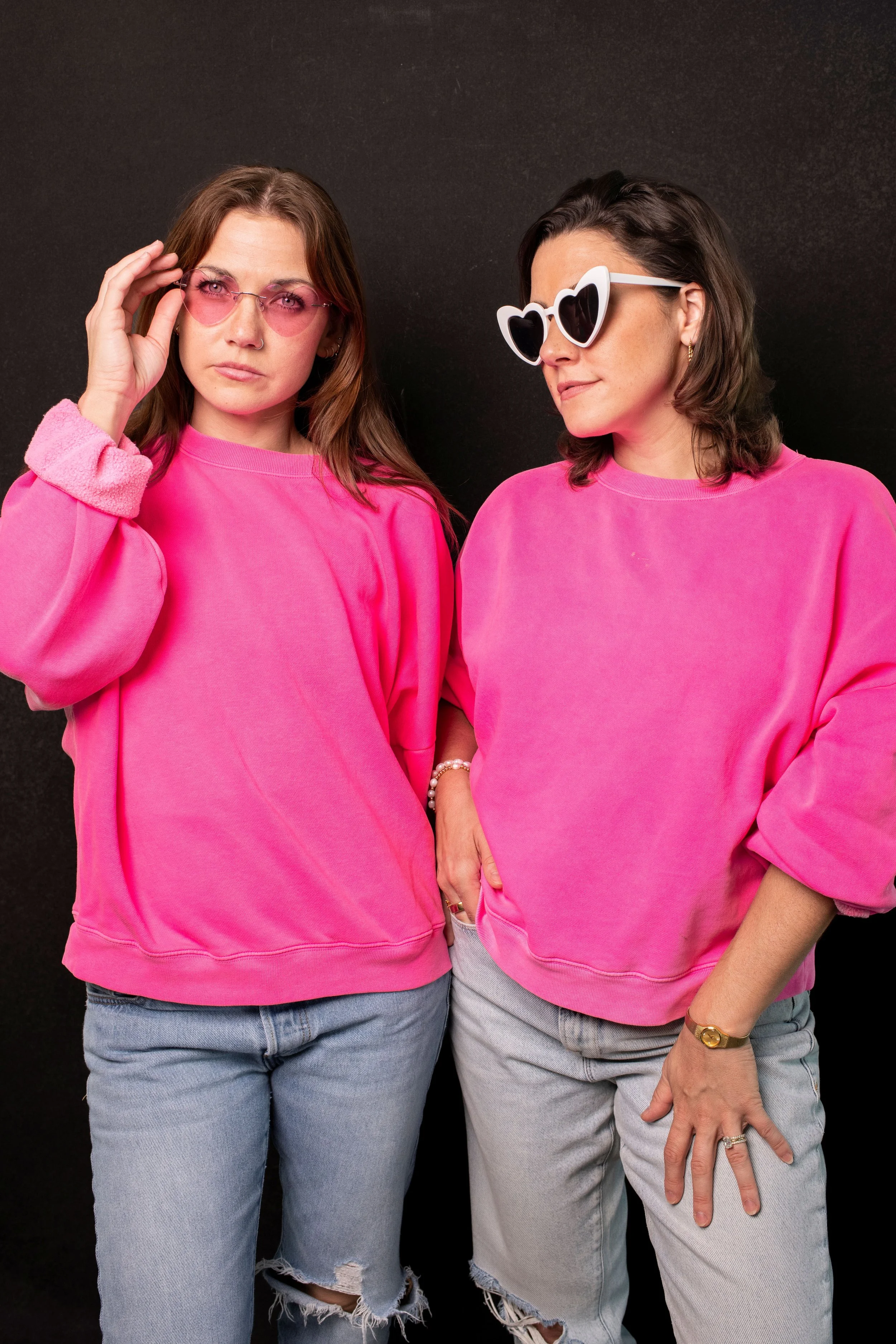 Two women wearing matching pink sweatshirts and jeans, standing against a black background, with one adjusting her pink-tinted glasses and the other wearing heart-shaped sunglasses.