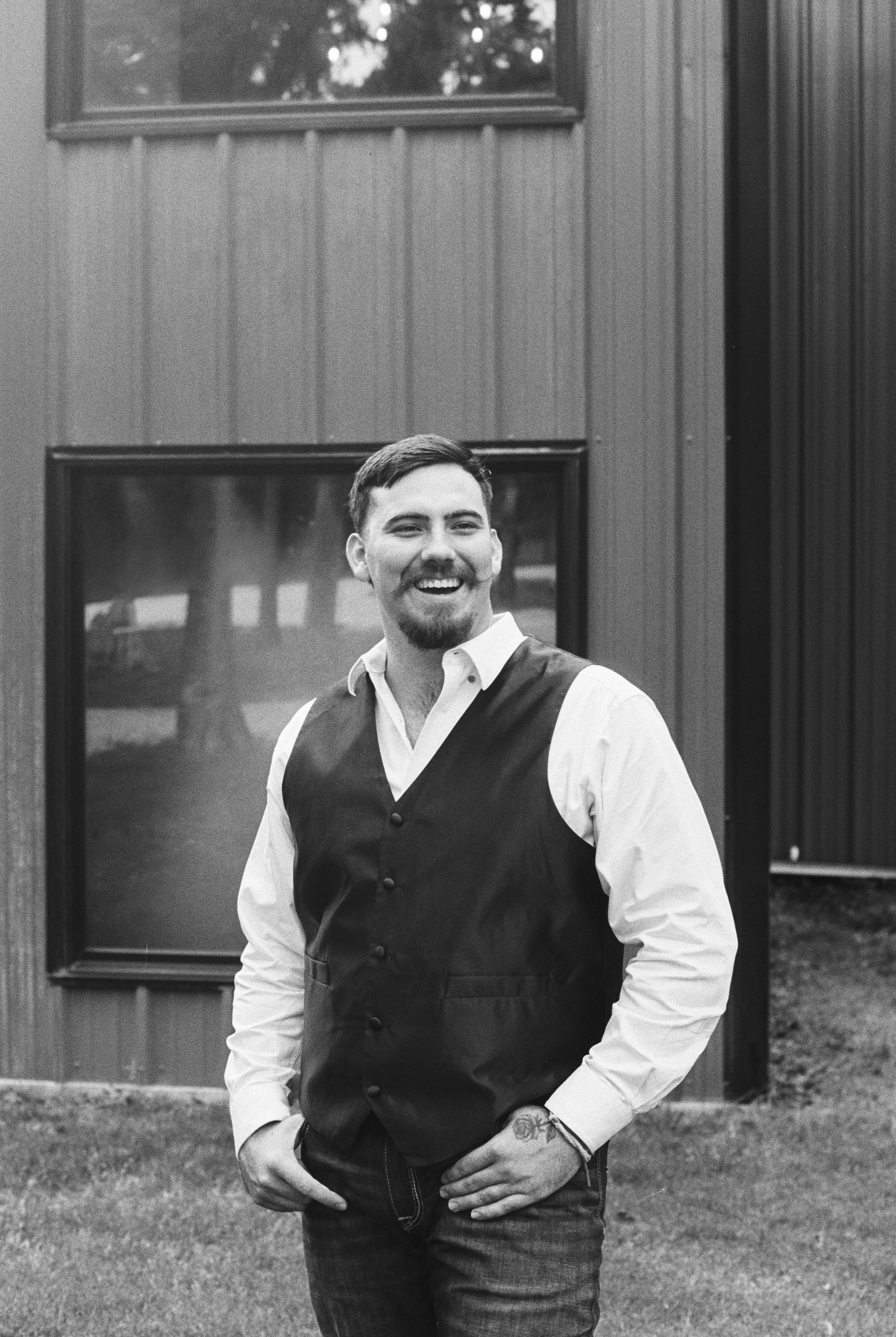 A smiling man with a beard and mustache, wearing a white shirt and a dark vest, standing outdoors in front of a building with wooden paneling and windows, with trees reflected in the window.