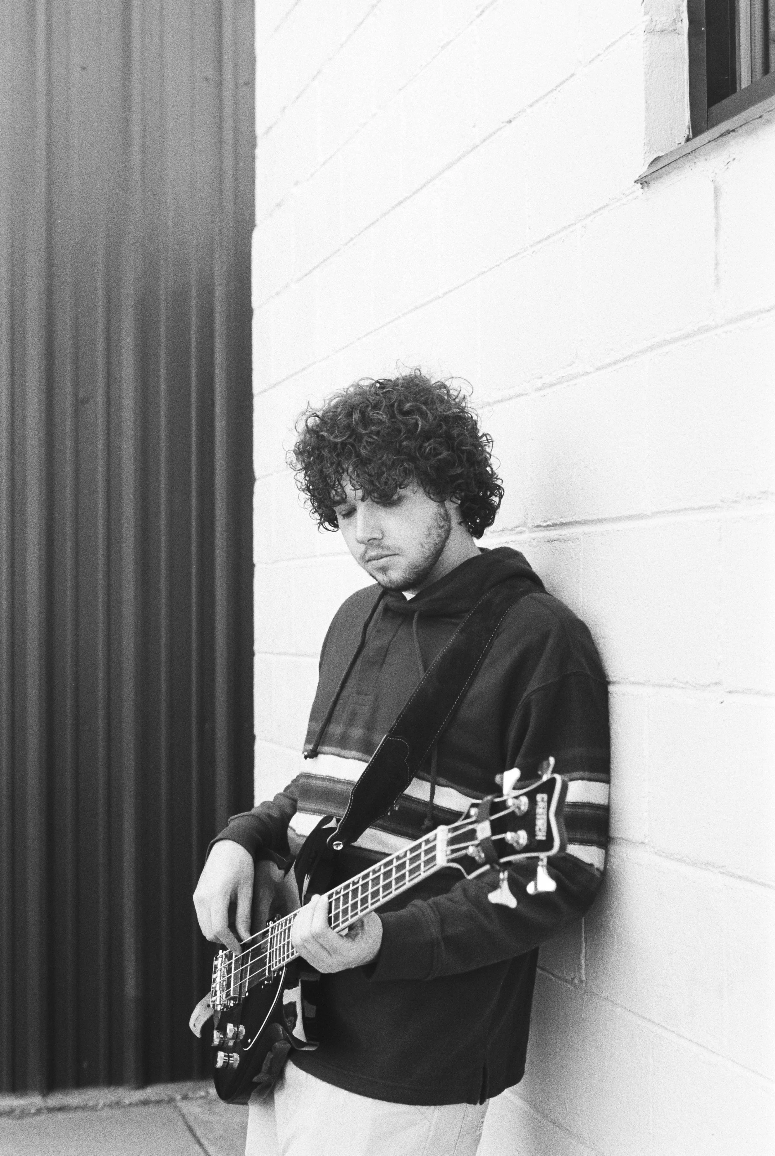 A young man with curly hair standing against a white brick wall, playing an electric bass guitar.