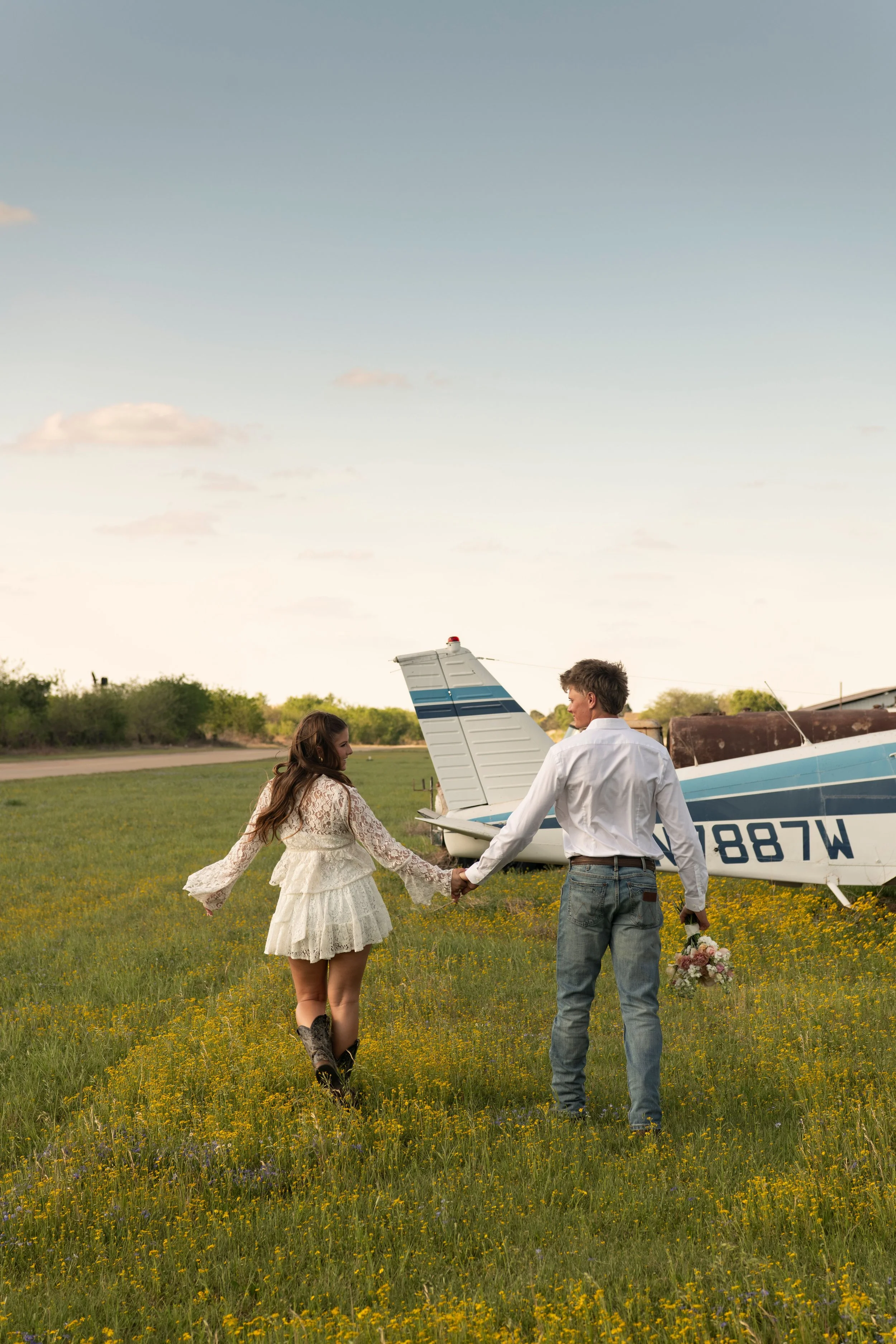 A man and woman holding hands in a field with yellow flowers near small airplanes parked in the background under a partly cloudy sky.