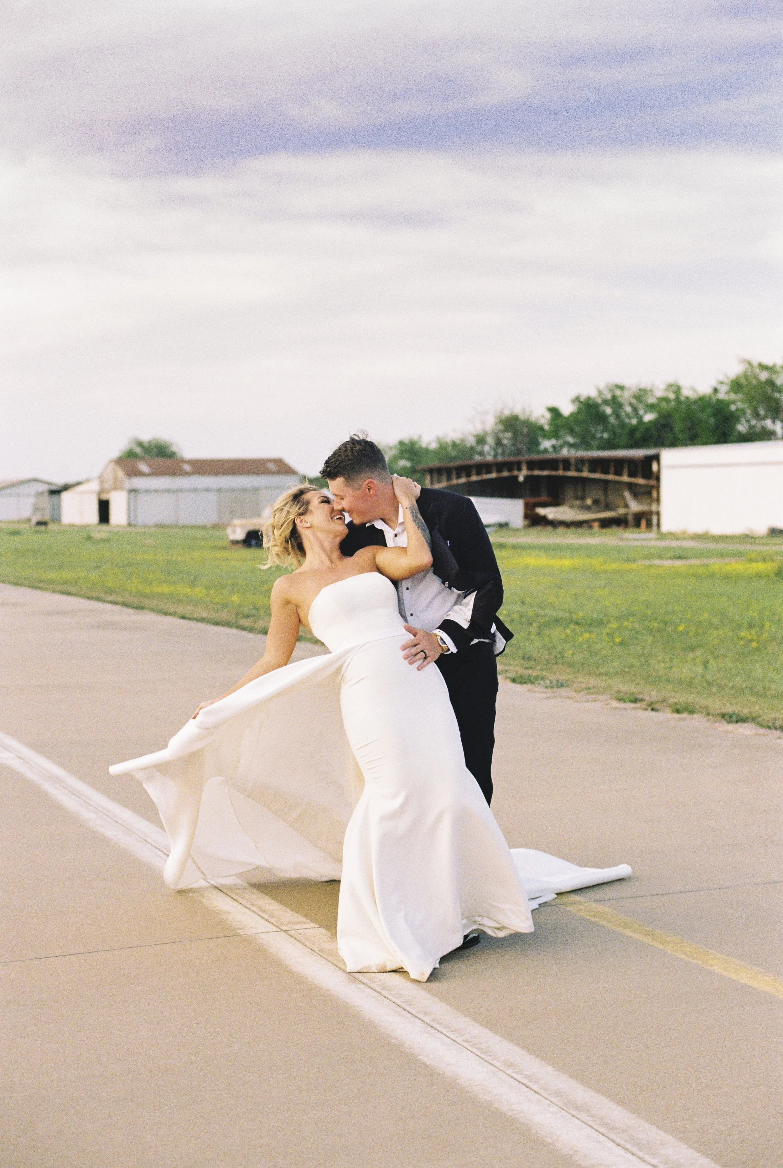 A newlywed couple dancing on an empty outdoor runway or road, with the bride in a white wedding dress and the groom in a black suit, smiling and embracing each other.