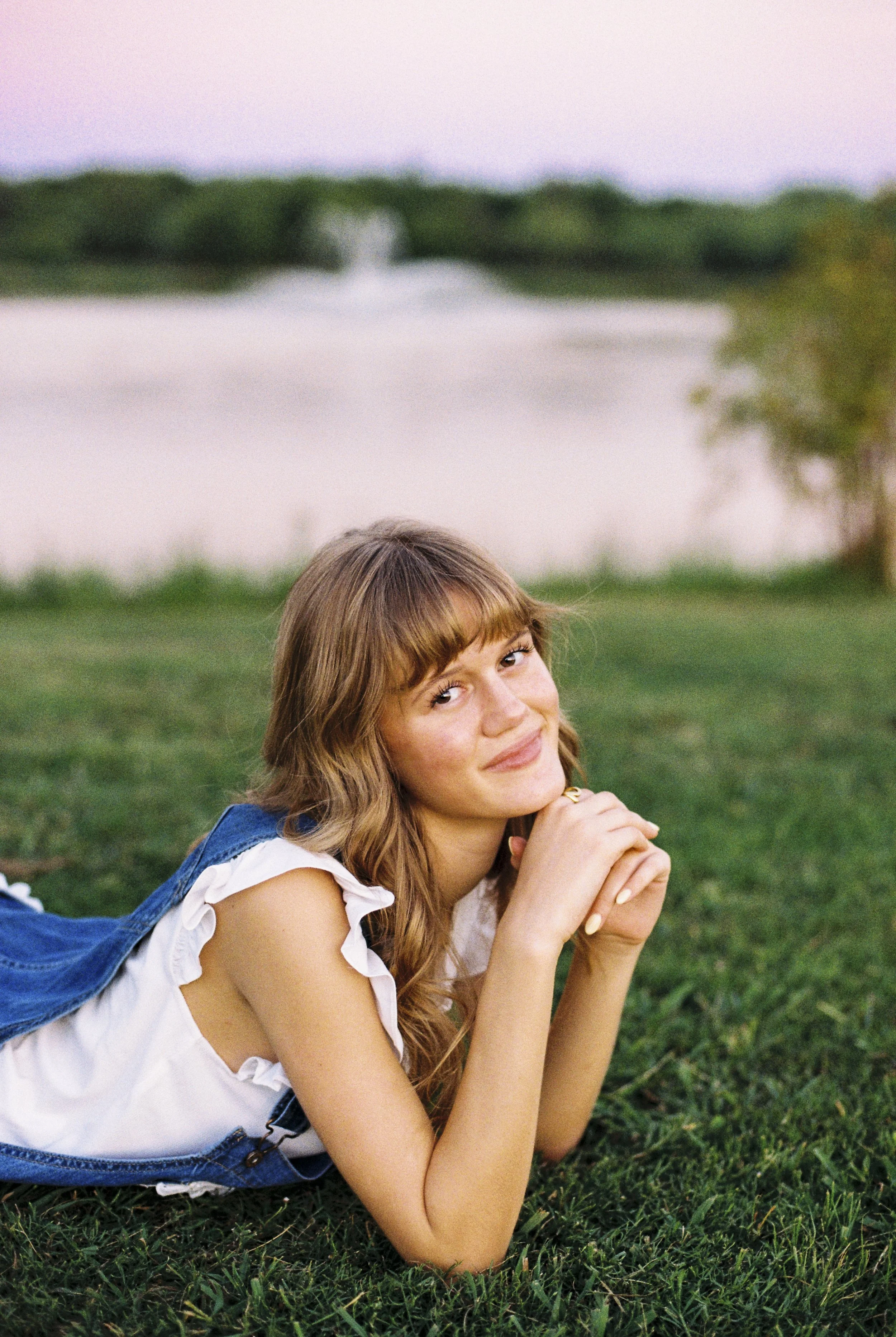 A young woman with long wavy hair, lying on the grass by a lake, smiling at the camera during sunset.