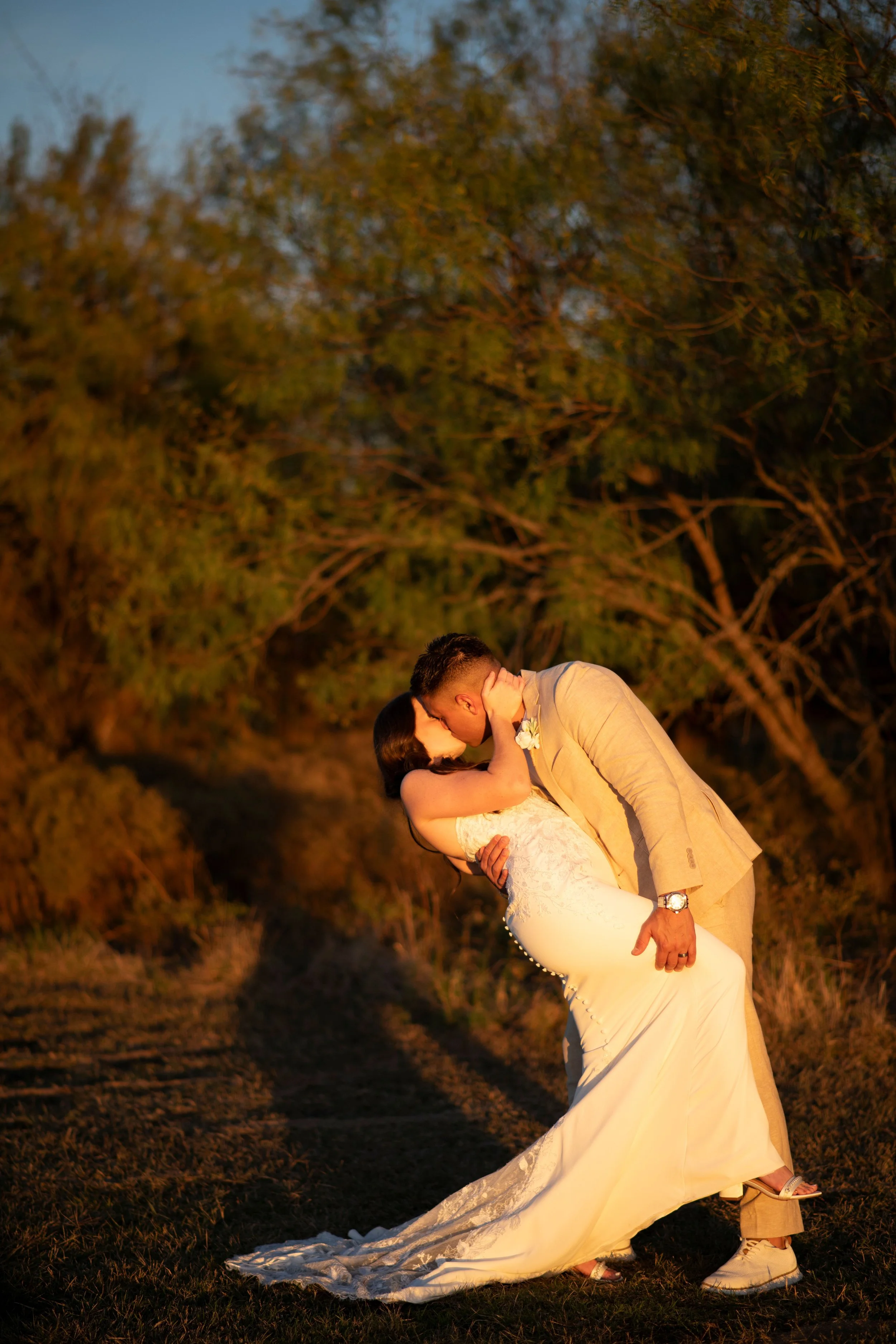 A couple in wedding attire sharing a kiss outdoors during sunset, surrounded by trees and natural scenery.
