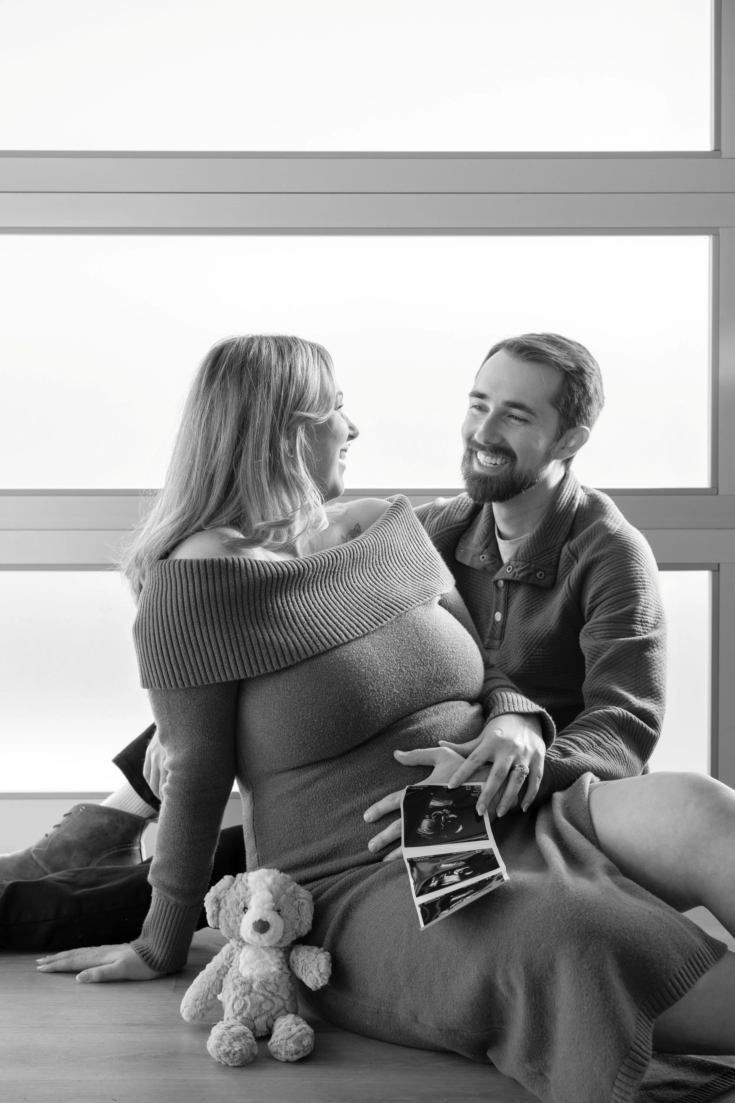 Black and white photo of a pregnant woman and her partner smiling at each other, sitting on the floor with the woman's ultrasound photos and a stuffed animal nearby, in front of large windows.