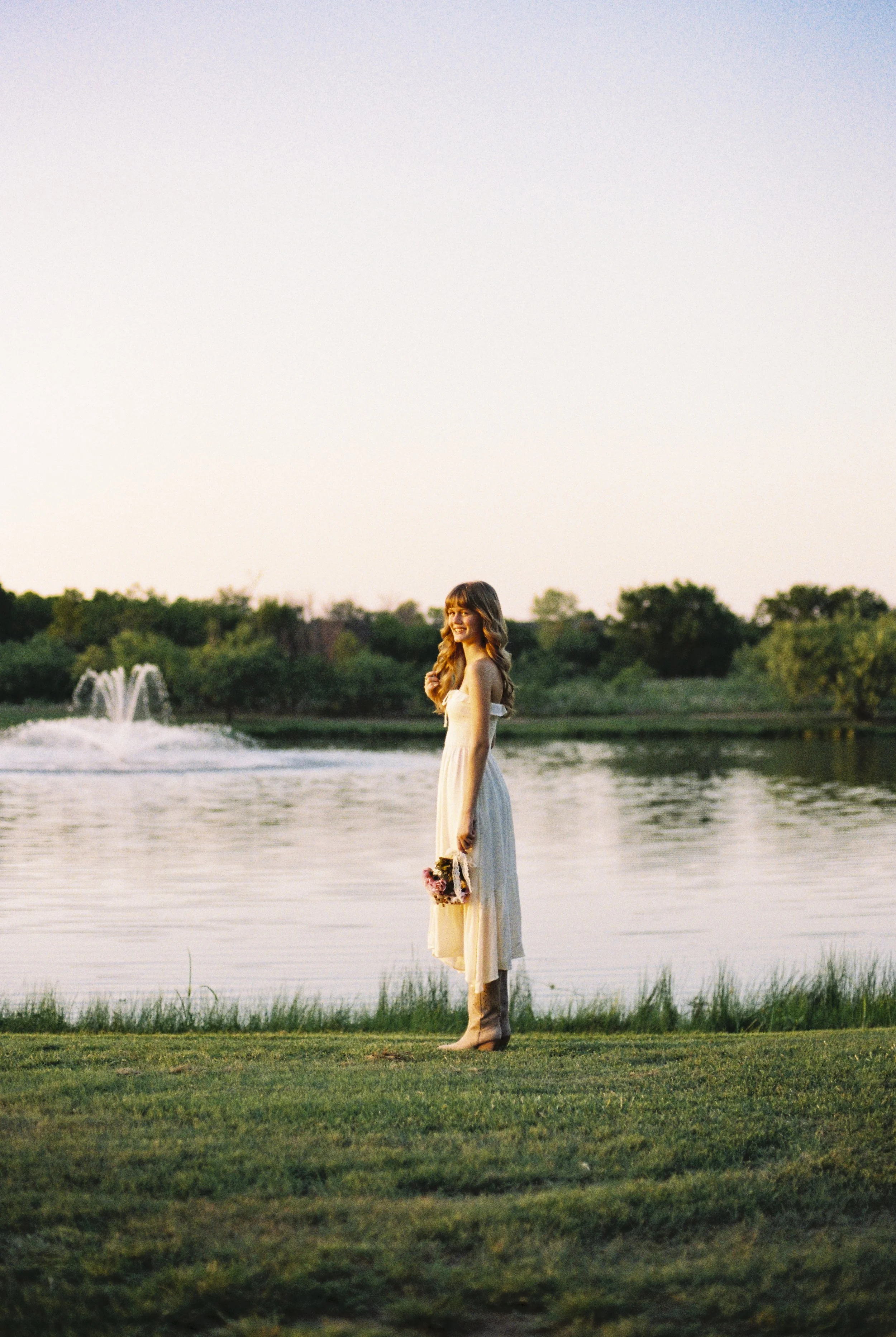 A teen in a yellow dress holding a bouquet stands near a lake during sunset, with a fountain in the background and lush greenery around.