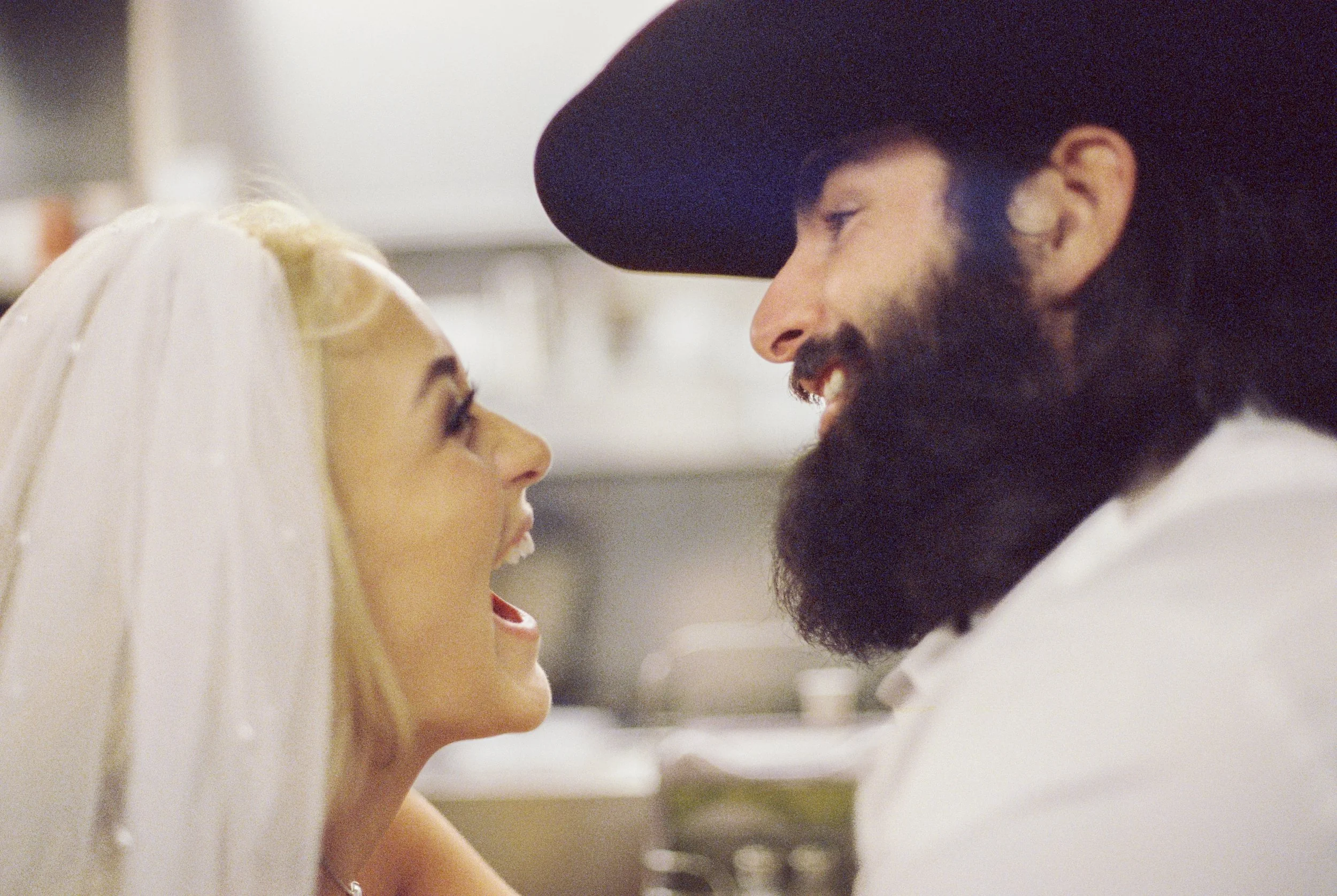 A bride and groom face each other smiling in a close-up shot with a blurry background.