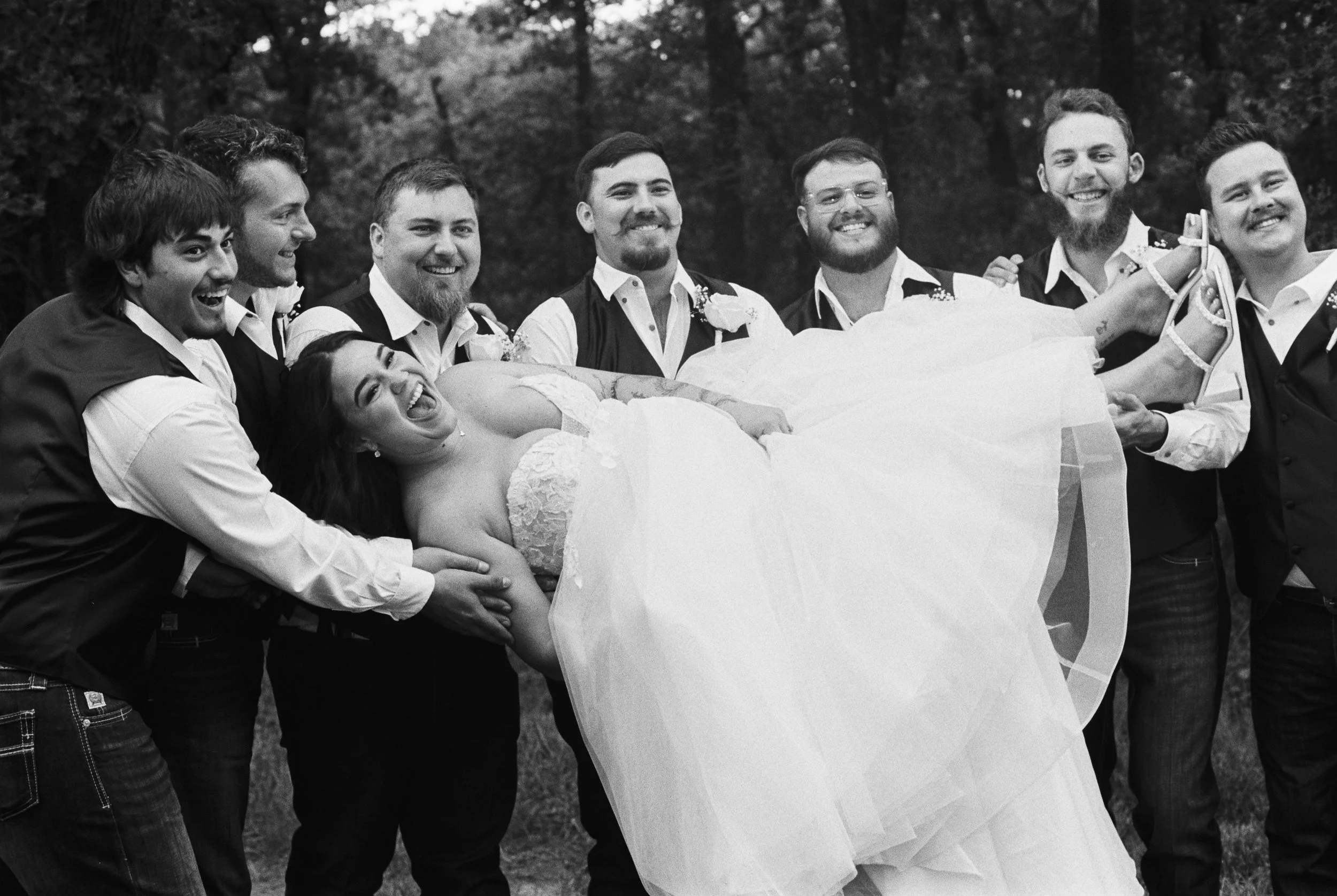 A group of men in formal attire lifting a woman in a wedding dress during an outdoor wedding celebration.