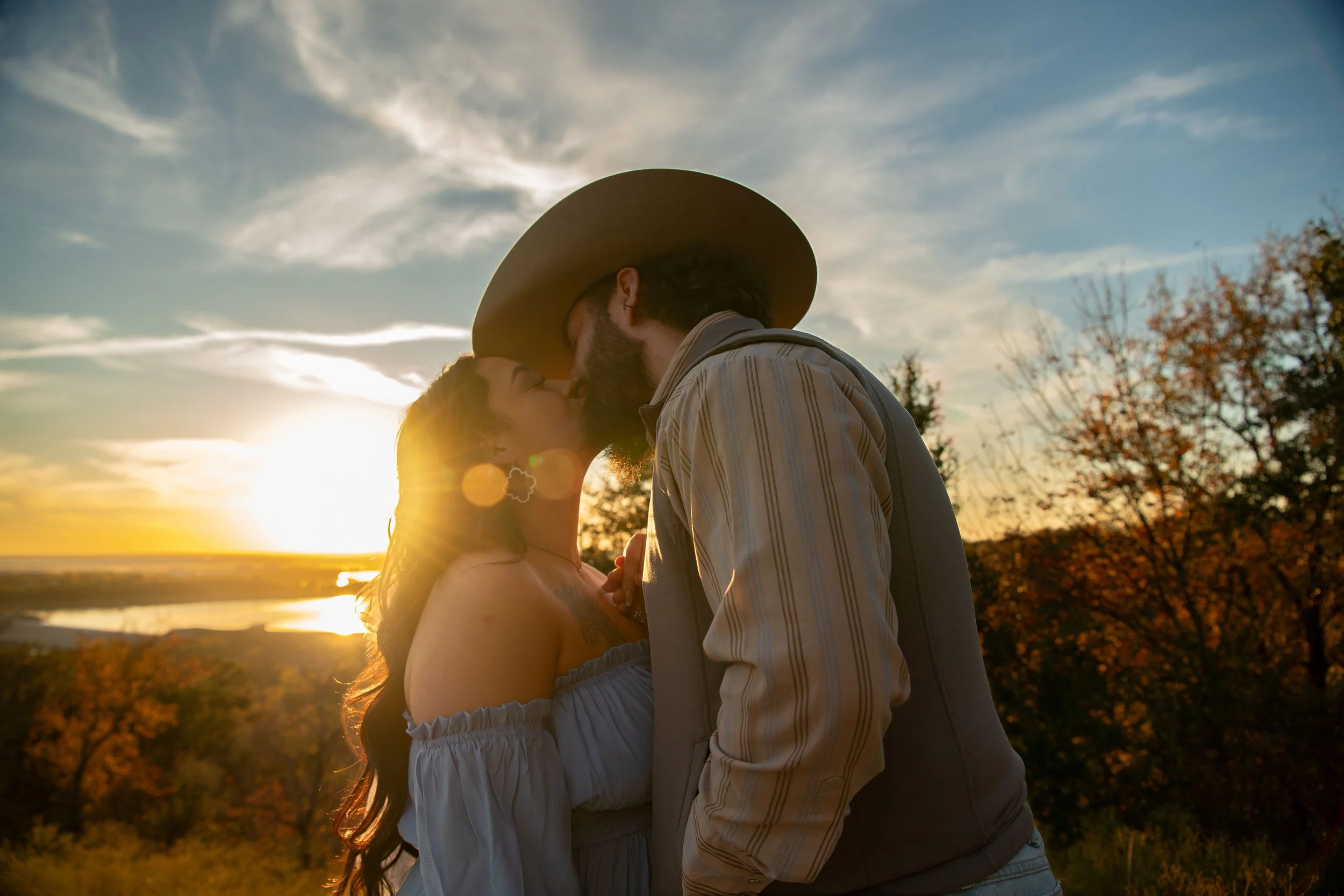 A couple sharing a kiss outdoors during sunset, with trees and a river in the background.