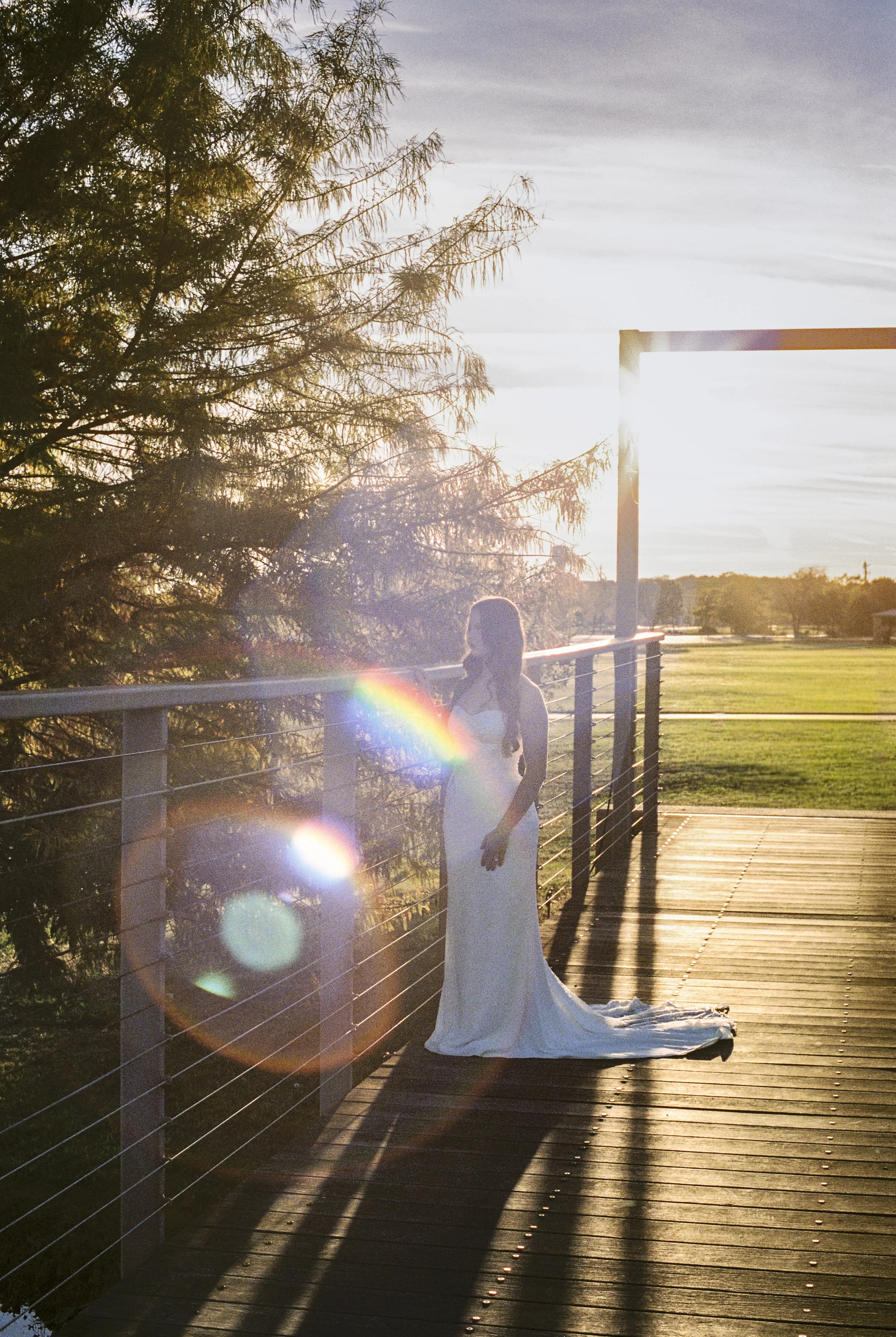 A woman in a white wedding dress stands on a wooden bridge at sunset, casting a long shadow and surrounded by lens flare effects.