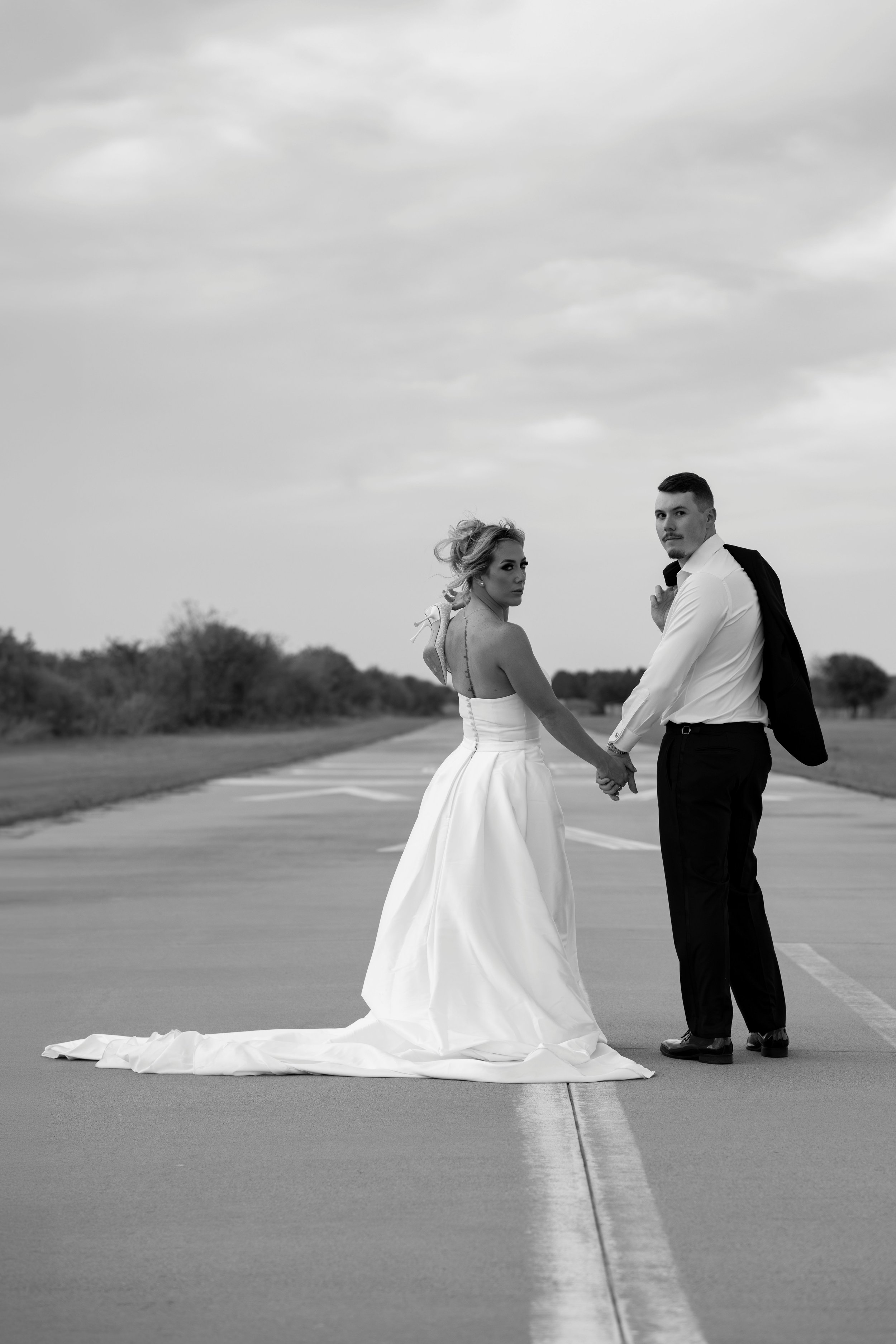 A black and white photo of a bride and groom holding hands on an empty runway outdoors. The bride wears a strapless wedding gown, and the groom wears a white shirt with black pants and a suit jacket draped over his shoulder. The sky is cloudy, and th