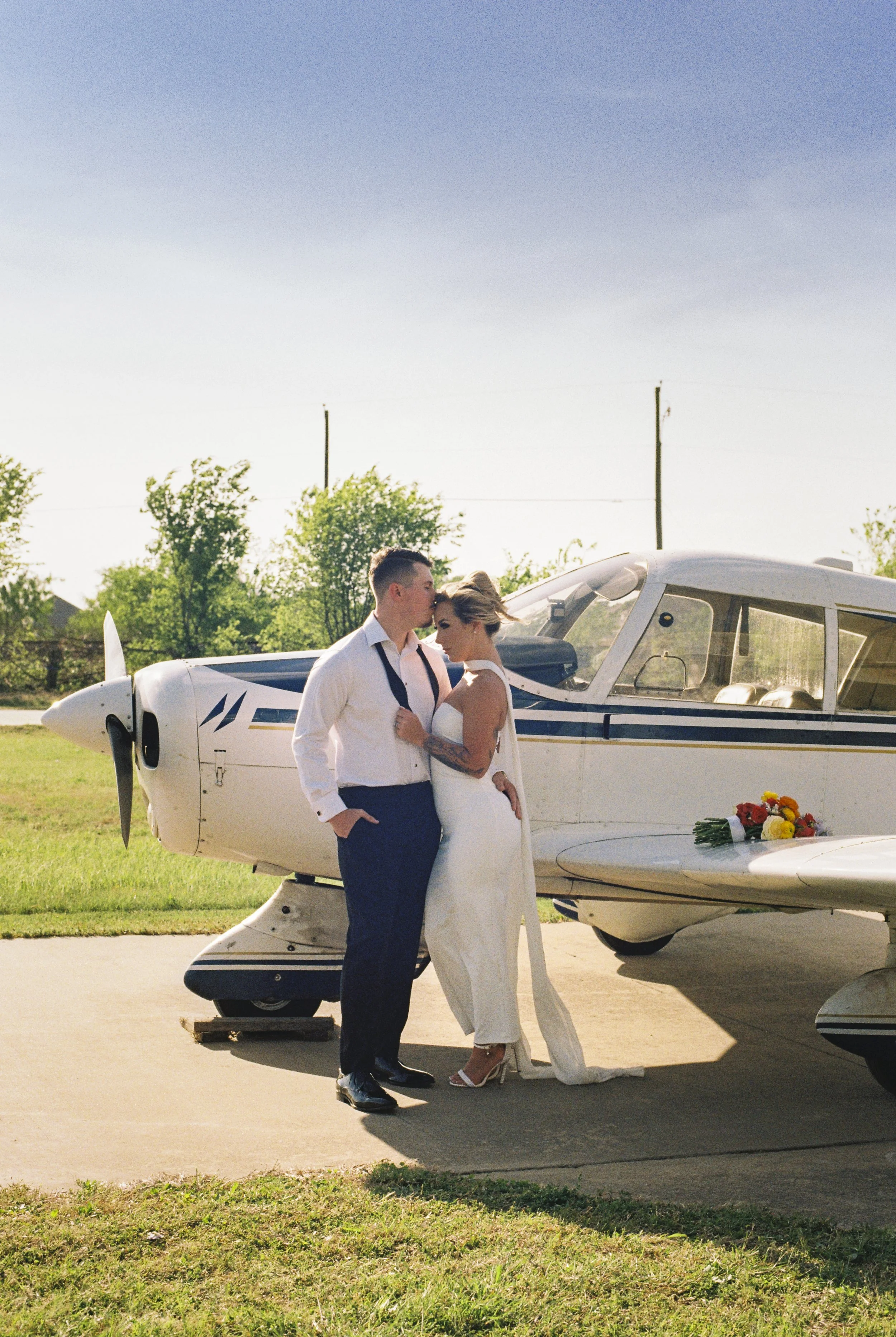 A couple dressed in wedding attire standing close to each other in front of a small airplane, with a bouquet of flowers placed on the airplane's wing, outdoors on a sunny day.