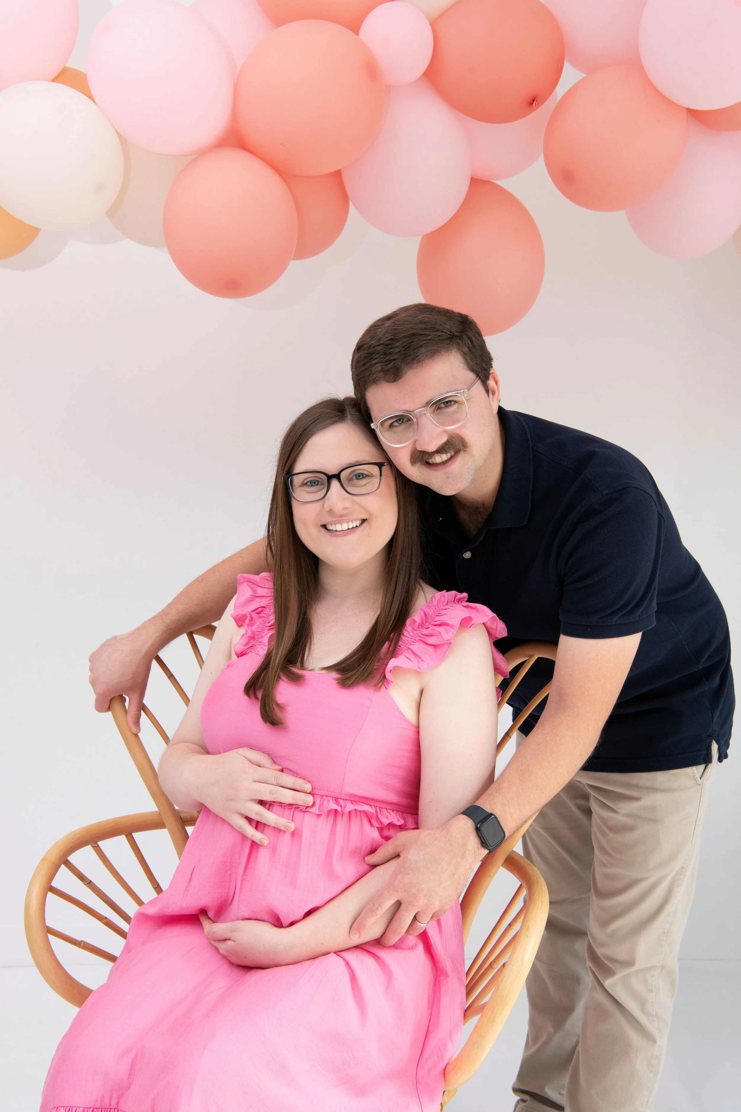 A pregnant woman in a pink dress sitting on a wooden chair, smiling, with a man standing beside her, both smiling, against a backdrop of pink and white balloons.