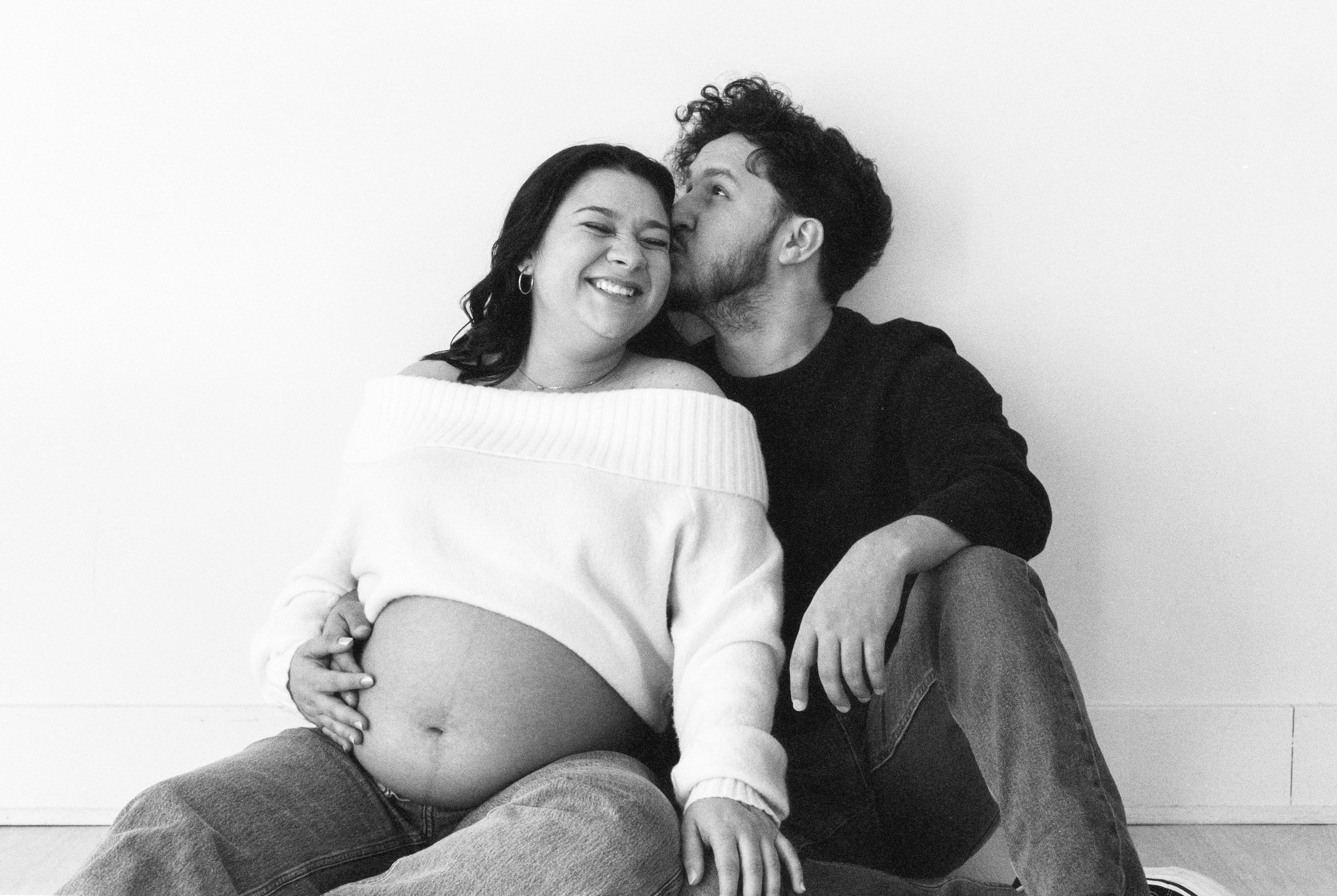 A pregnant woman with dark hair smiling, sitting on the floor, with a man with curly hair and beard kissing her cheek, both sitting against a plain white wall.