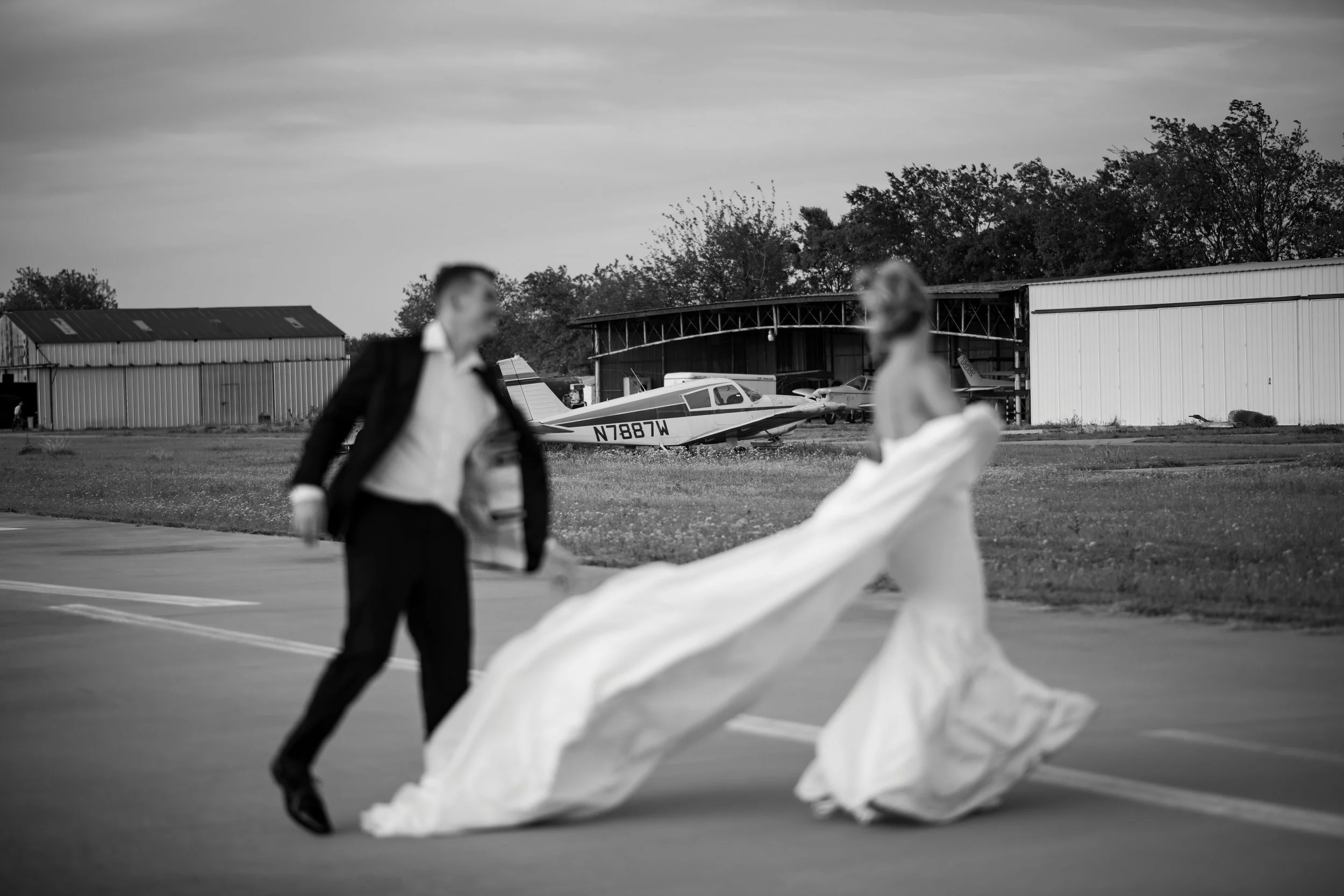 People in formal attire standing on an airfield with small airplanes in the background, one of which has the registration N7887W.