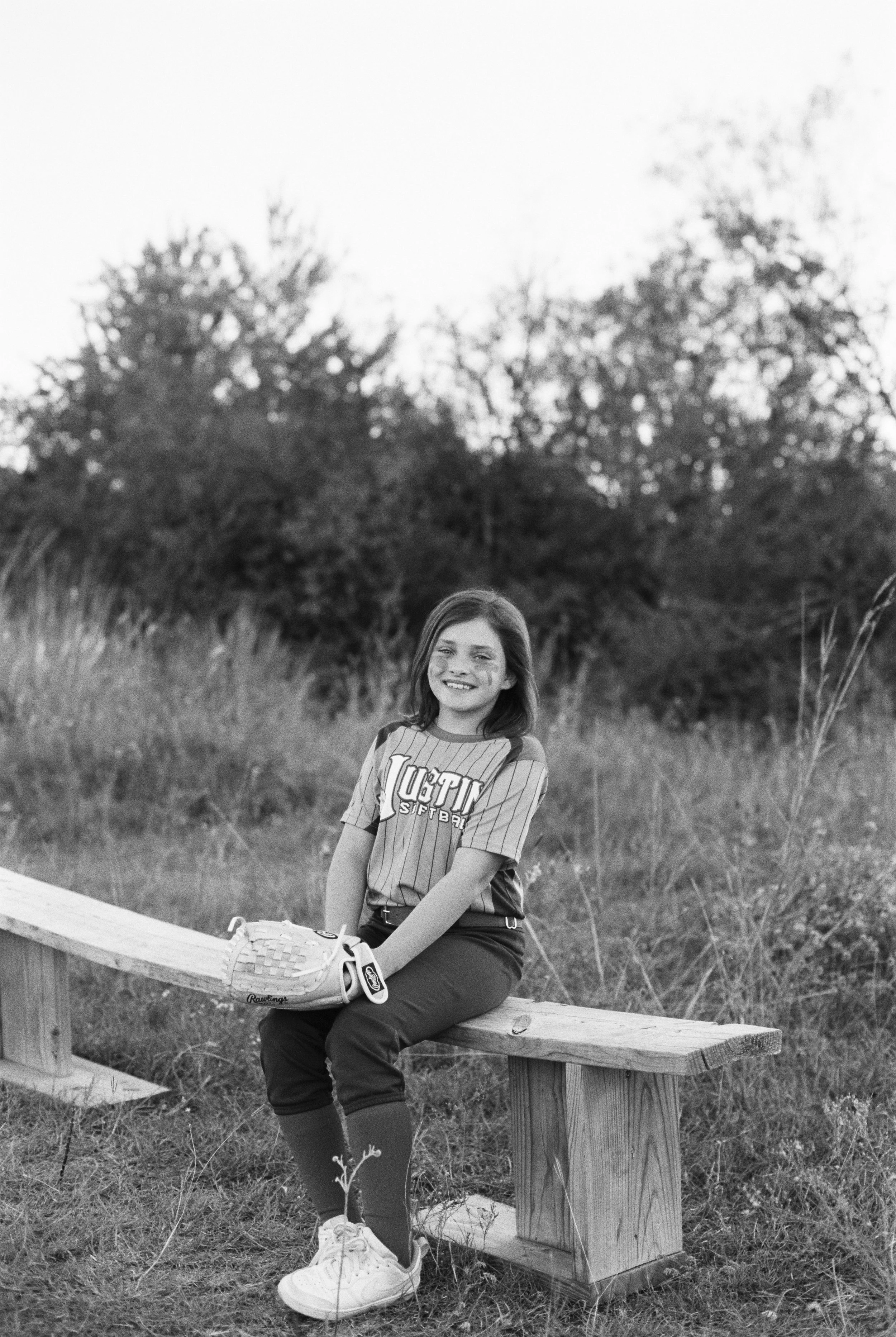 A girl in softball uniform sitting on a wooden bench outdoors, holding a softball glove, smiling at the camera, with trees in the background.