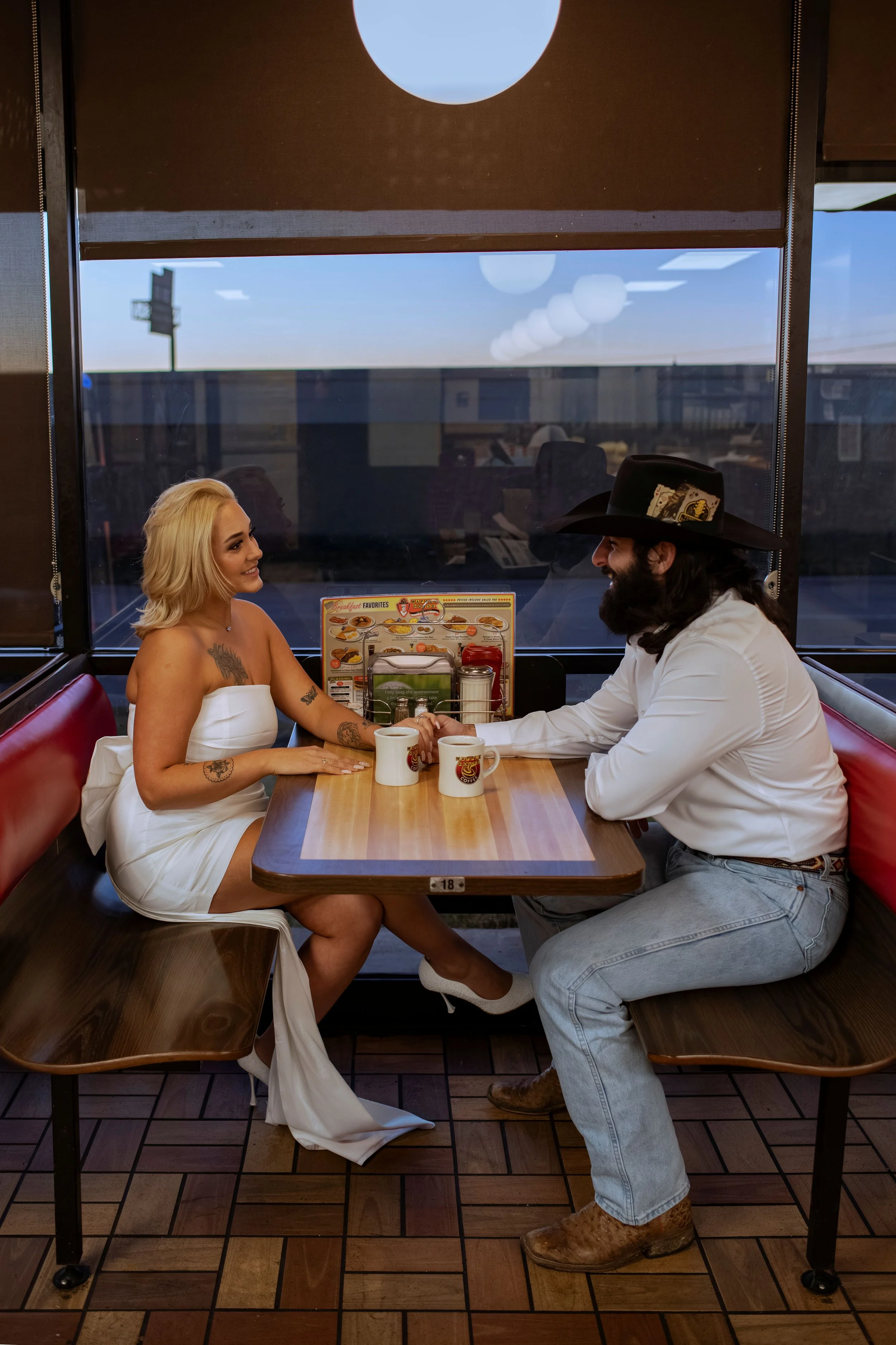 A woman with blonde hair, tattoos, and a white dress holding hands with a man wearing a large black hat, white shirt, gray pants, and brown boots, seated at a restaurant table with coffee mugs, inside a restaurant with large windows and wooden floori