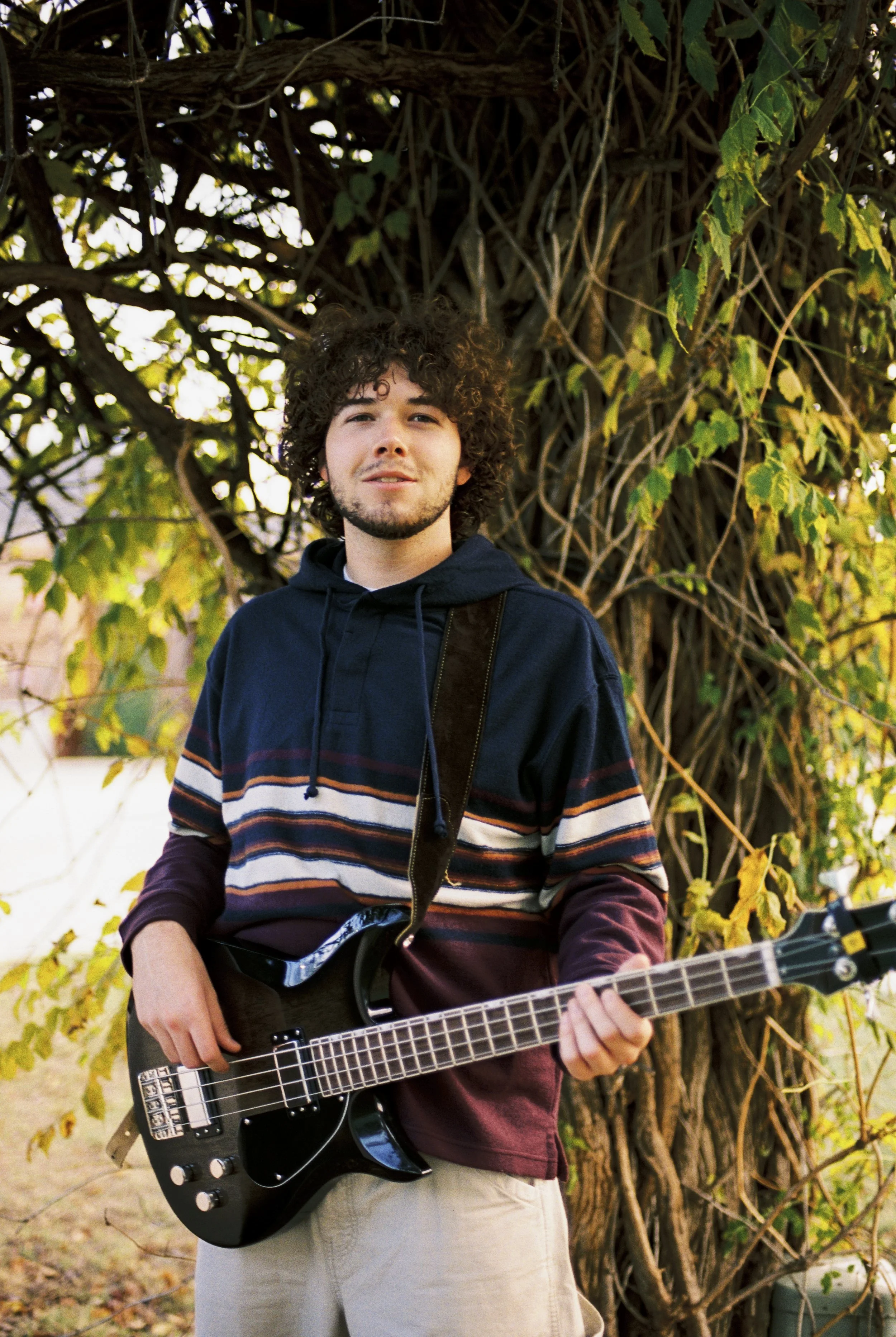 Young man with curly hair and facial hair holding a black electric bass guitar outdoors near a large tree with green and yellow leaves.