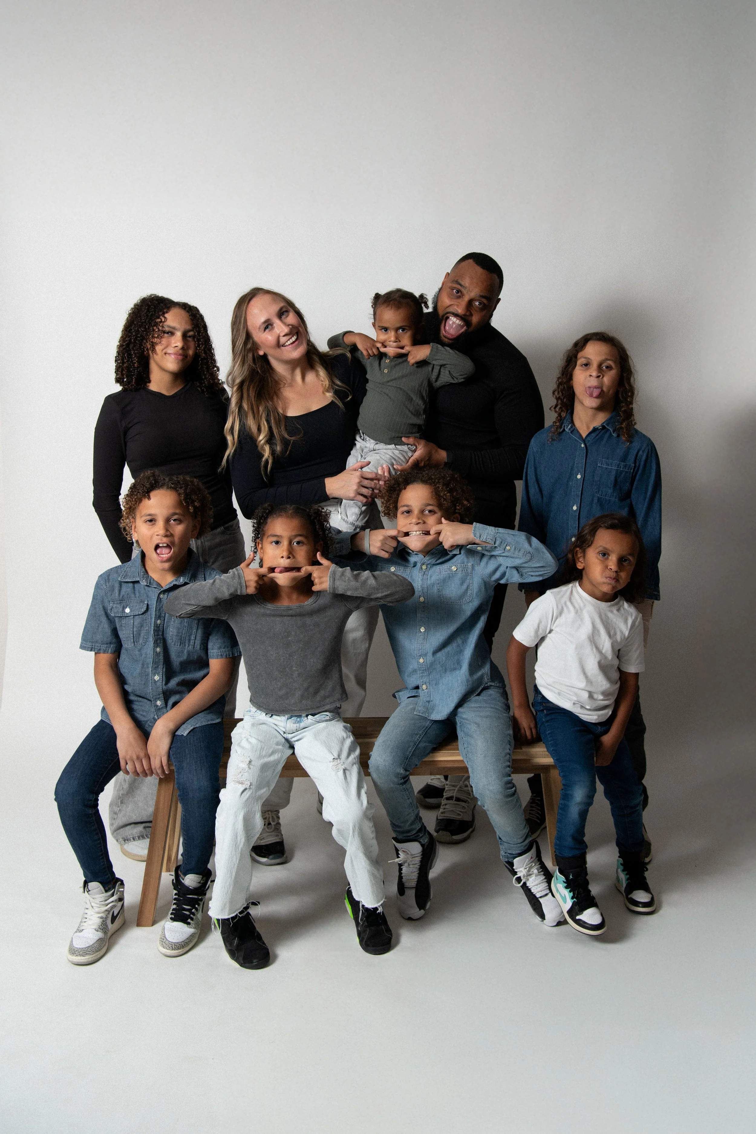 A diverse family of nine people posing playfully in front of a plain white background, some making funny faces.