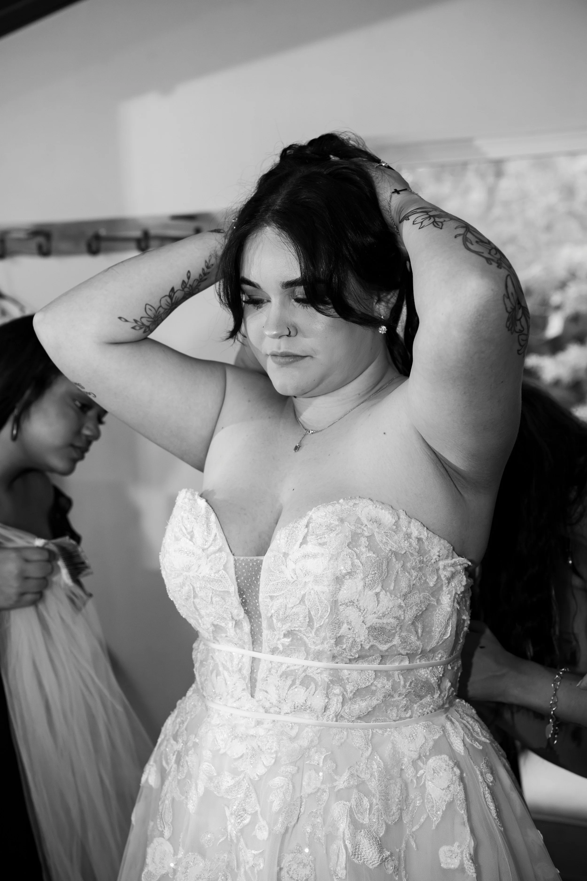 Black and white photograph of a bride adjusting her hair, wearing a strapless lace wedding gown with floral embroidery, and a necklace. Two women are in the background, one to the left and one to the right, amongst blurred indoor surroundings.