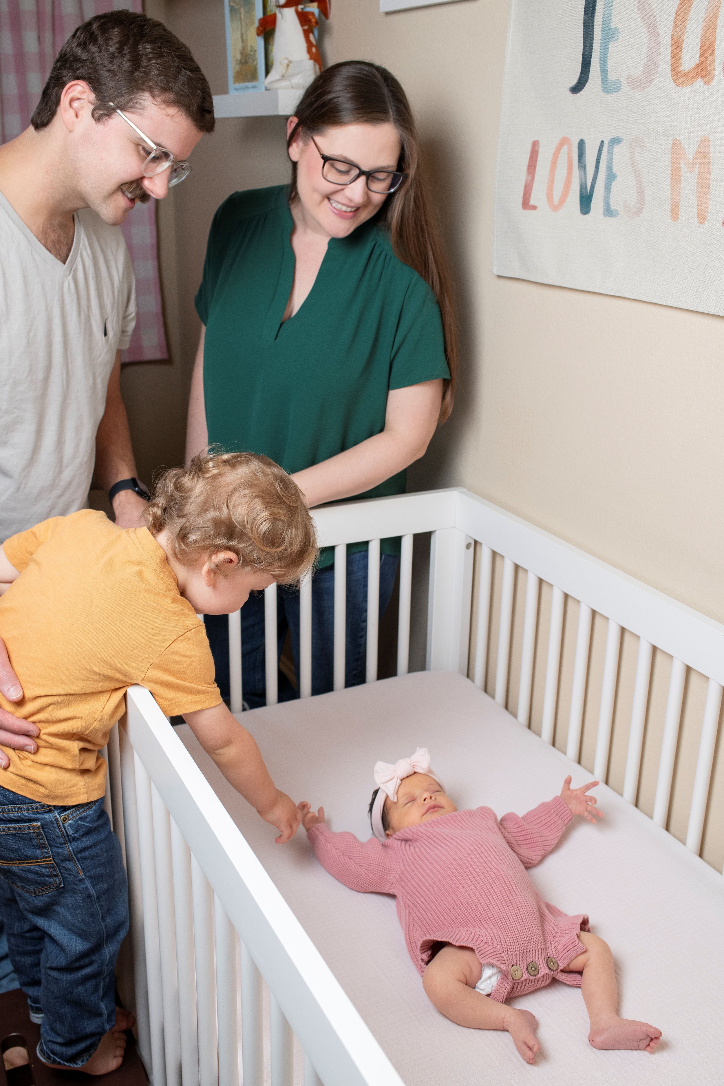 A family of four, including a baby girl in a pink outfit lying in a crib, a young boy in a yellow shirt reaching into the crib, a man with glasses wearing a beige shirt, and a woman with glasses and long brown hair in a green shirt, looking at the ba