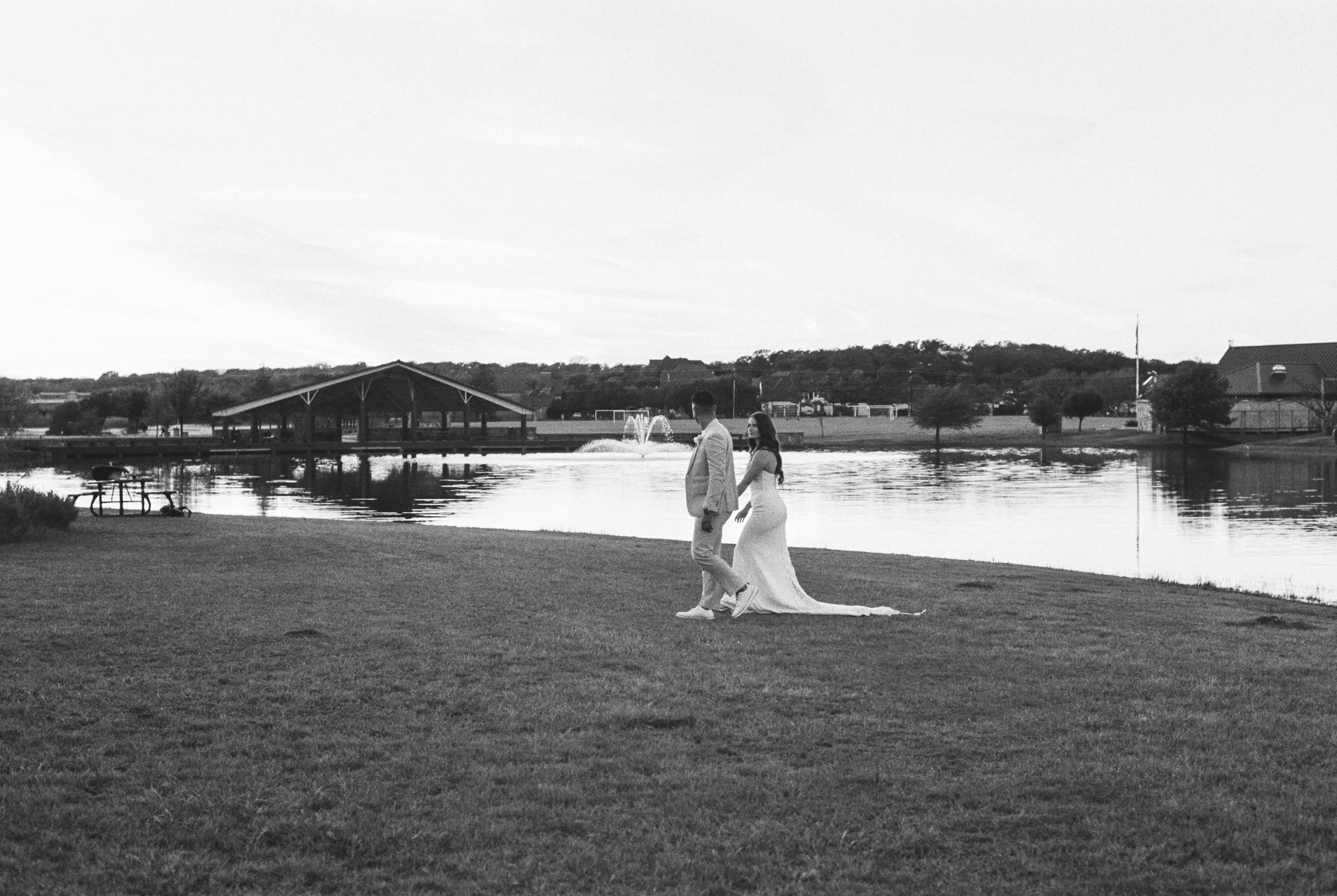 Black and white photo of a bride and groom holding hands, walking by a lake with a gazebo in the background.