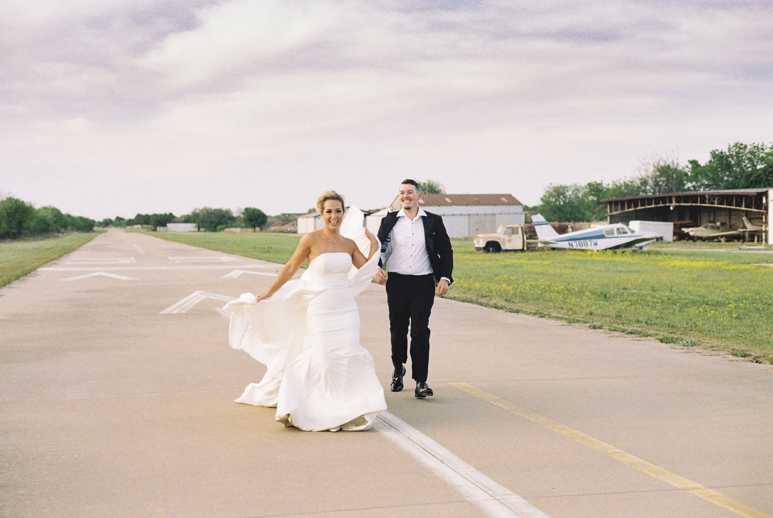 A bride and groom walking together on an empty runway at an airfield, with old airplanes and hangars in the background.