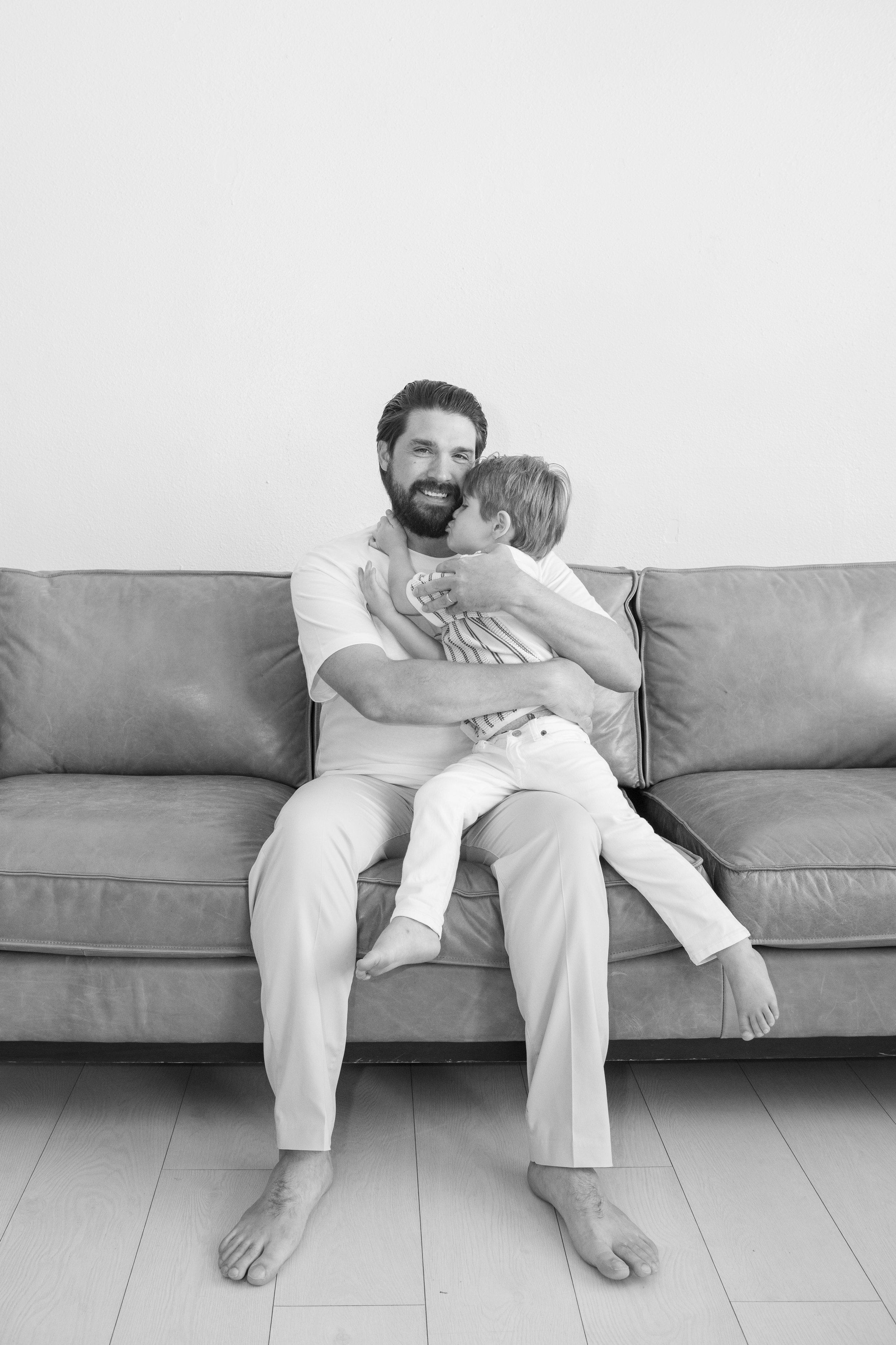 A man with a beard smiling while sitting on a sofa as he hugs and kisses a young child. The scene is in a minimalistic room with a plain wall behind them.