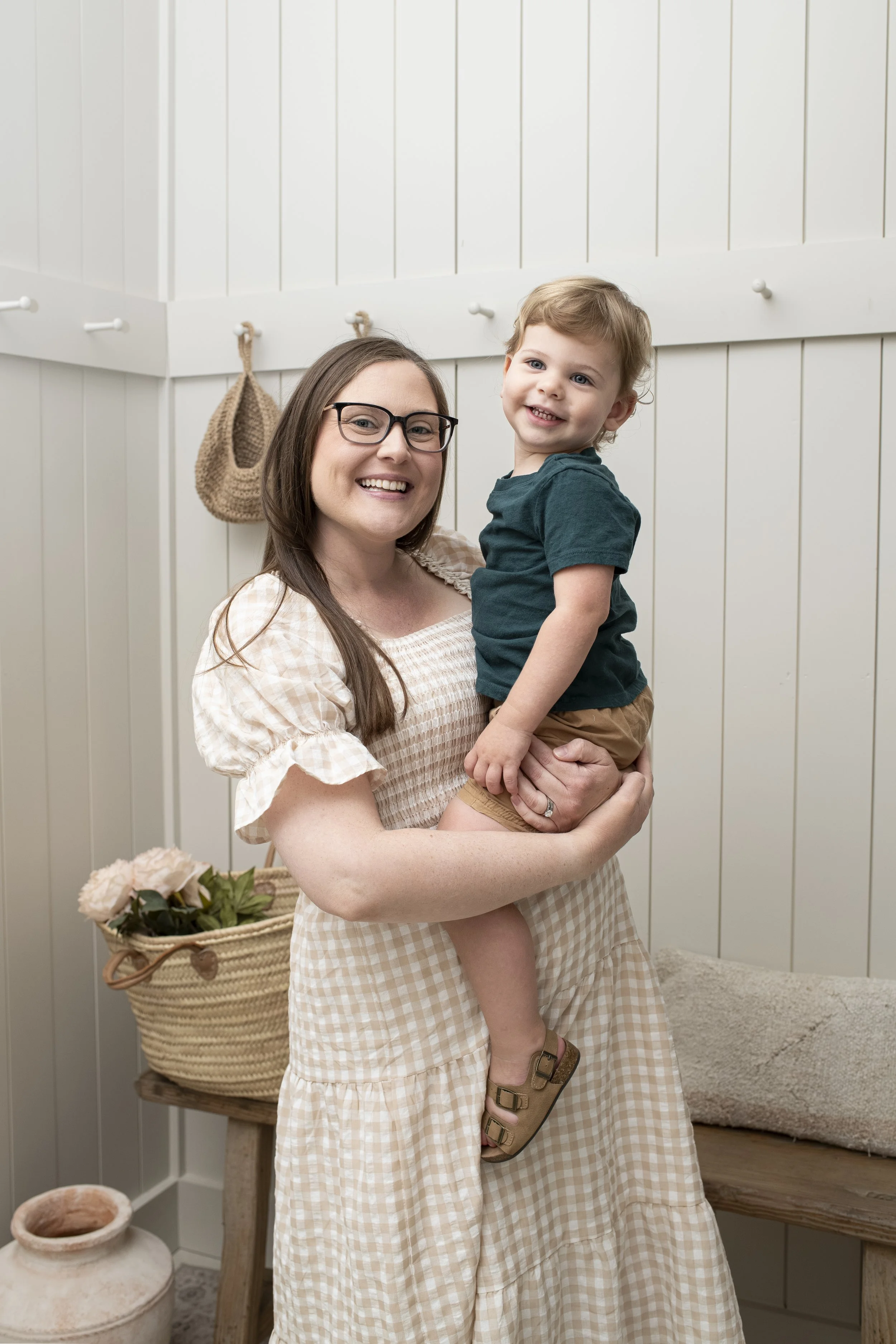 A woman with glasses and a beige checkered dress holding a young boy with curly hair in a green t-shirt and beige shorts, smiling indoors with white wooden panel walls, a basket of pink flowers, and a bench with a cushion.