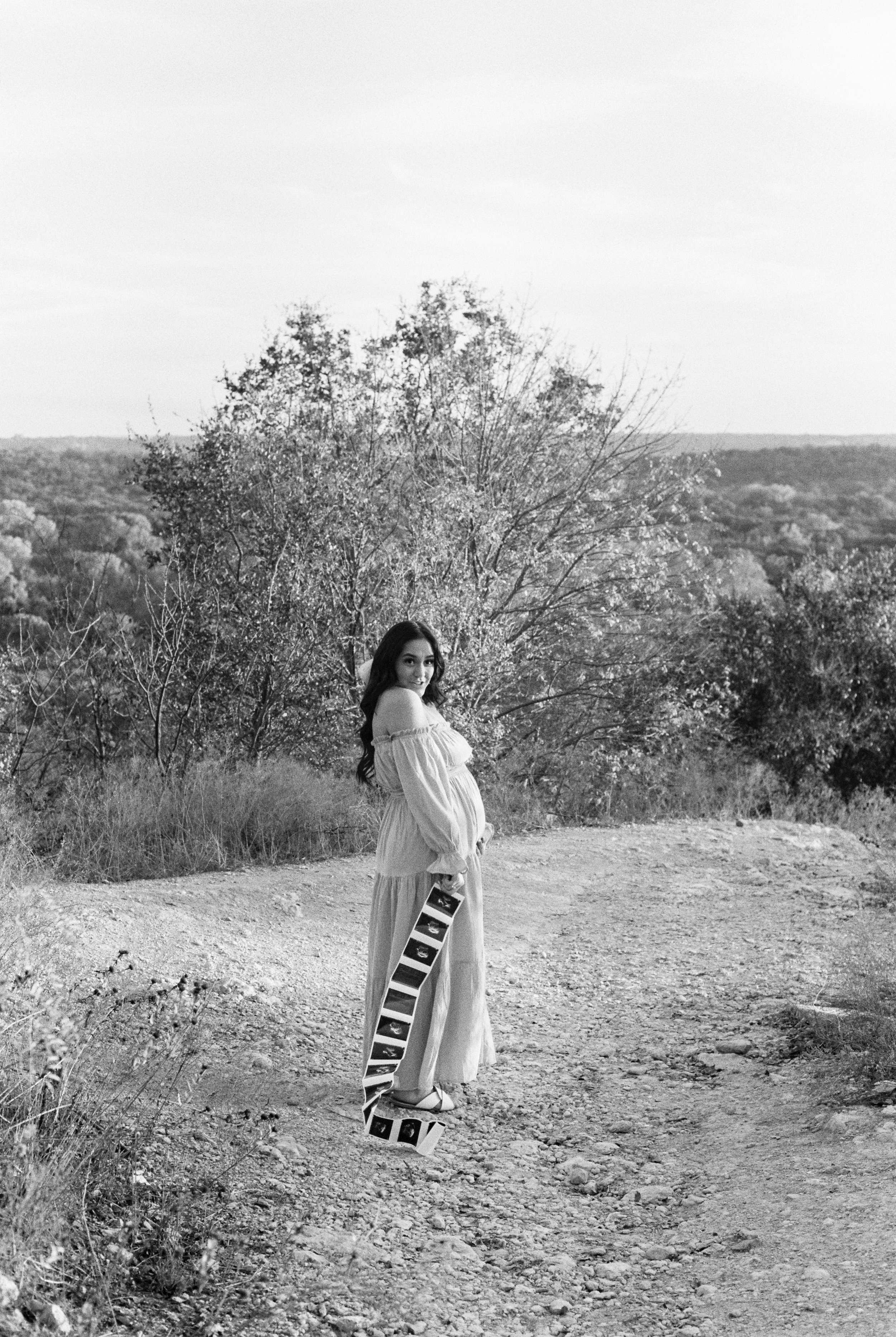A woman standing outdoors on a dirt path holding ultrasound images, with trees and a landscape in the background, in black and white.