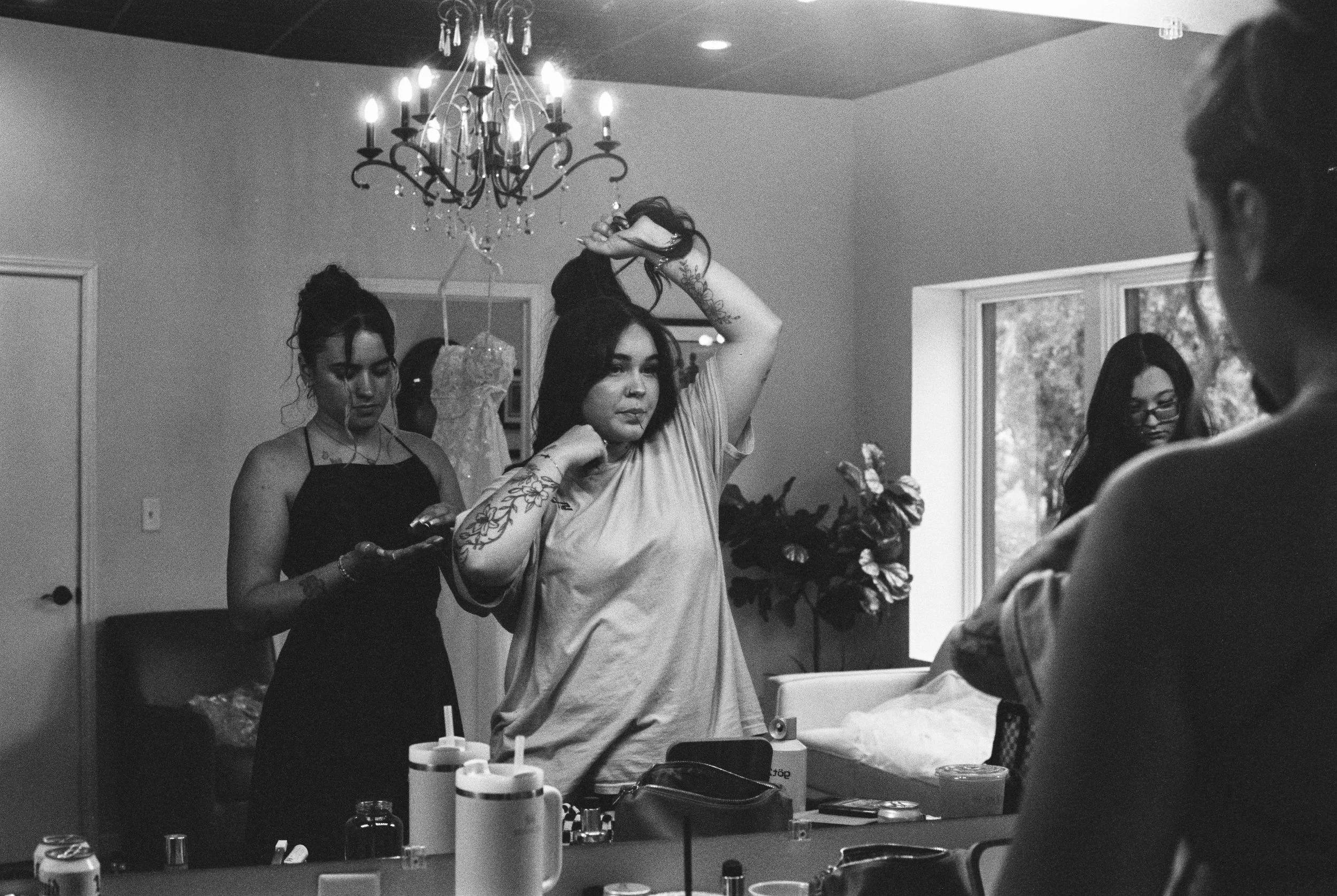 Women preparing or applying makeup or body art in a domestic setting with artistic tattoos, a chandelier, large window, and various toiletries and cosmetics on a table.